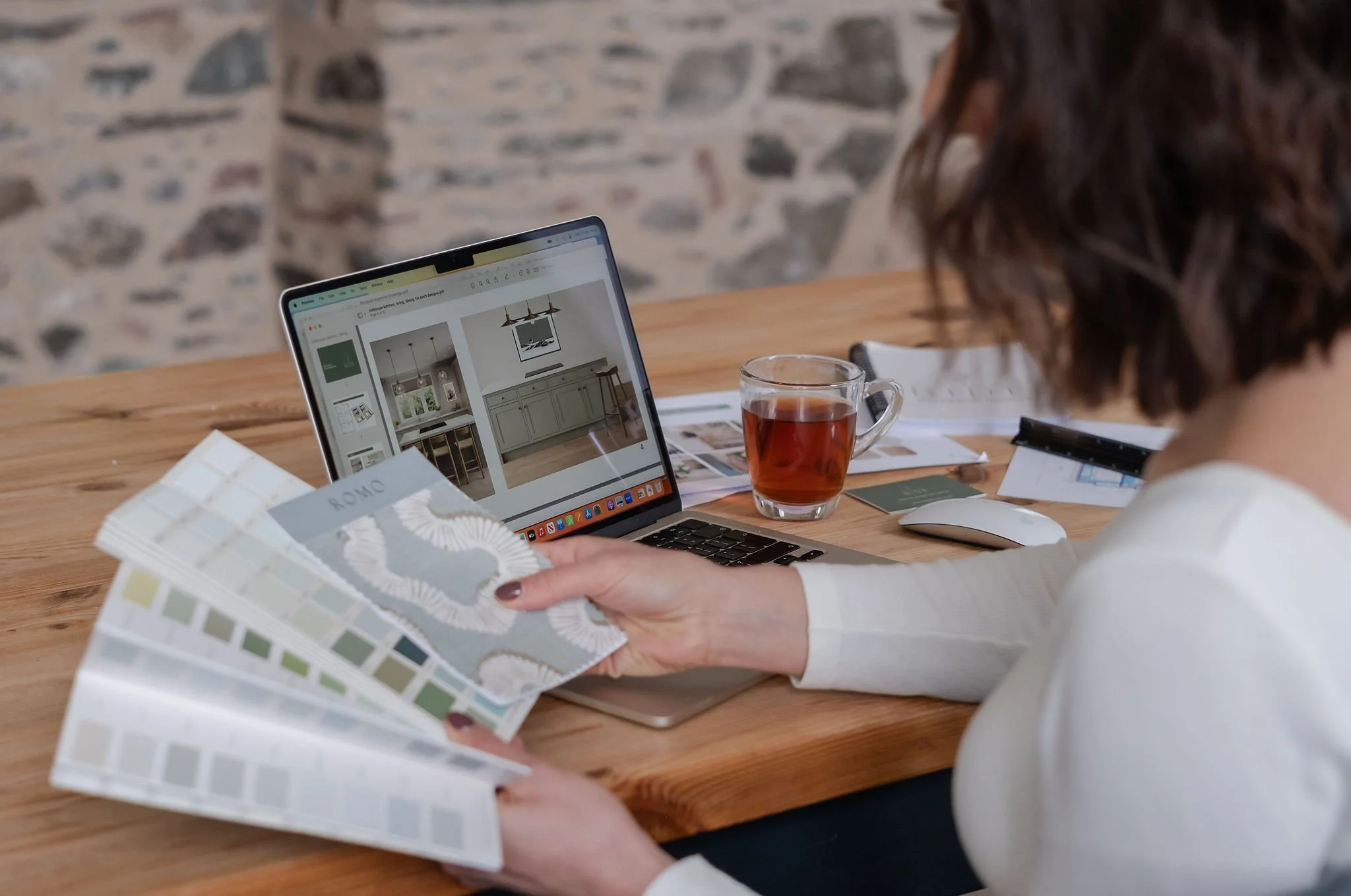 A person flips through fabric swatches and colour samples on a woven rug, with a basket containing additional fabric pieces and colour palettes nearby.