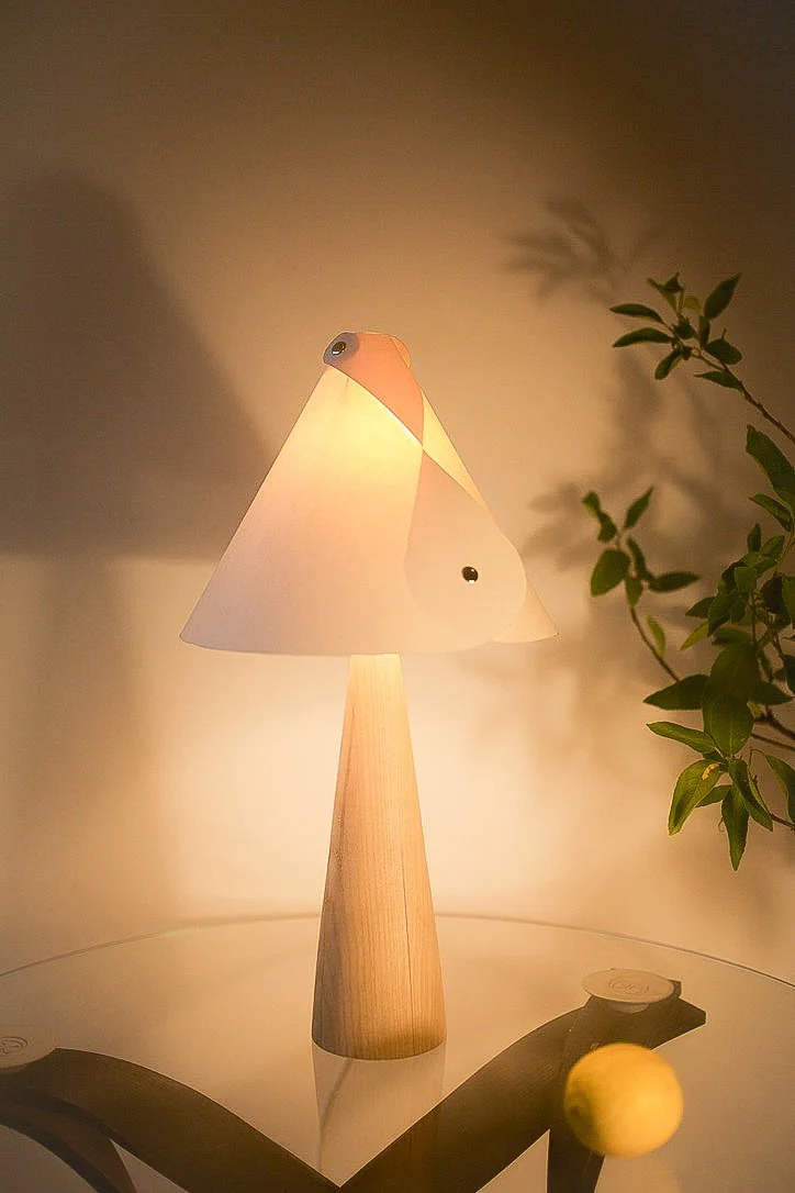 Table lamp with a bird-shaped lampshade and a wooden base, casting warm light in a room with a potted plant and yellow apple on a glass table.