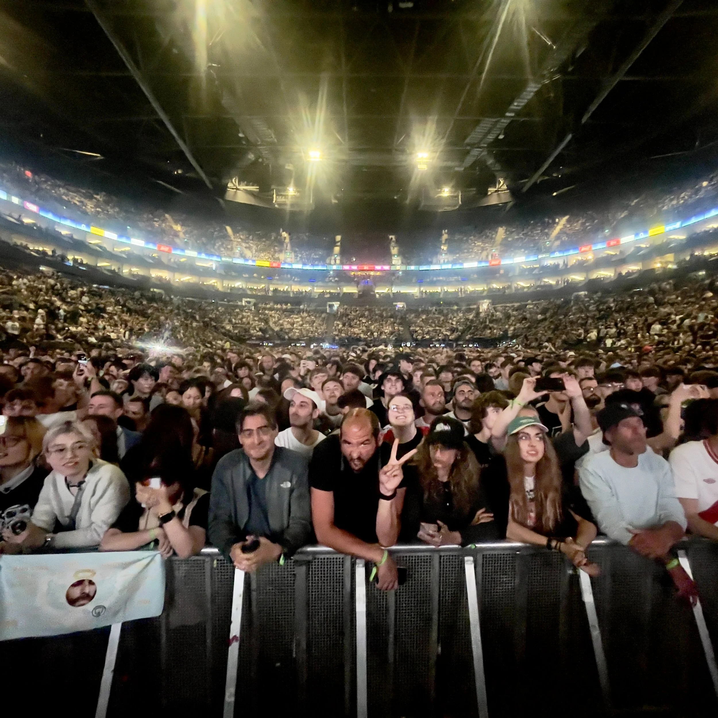 Crowd of people at a large indoor concert or event, with bright overhead lights and a stadium-style setting.