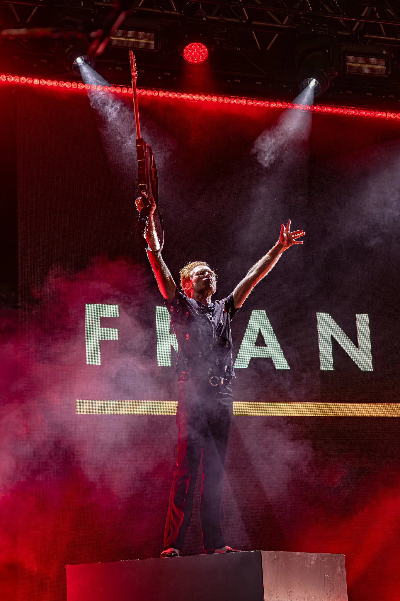 Performer standing on a stage with arms raised, holding a guitar in one hand. Stage is illuminated with red and white lights, with smoke effects surrounding the performer. A large screen behind displays part of a word.