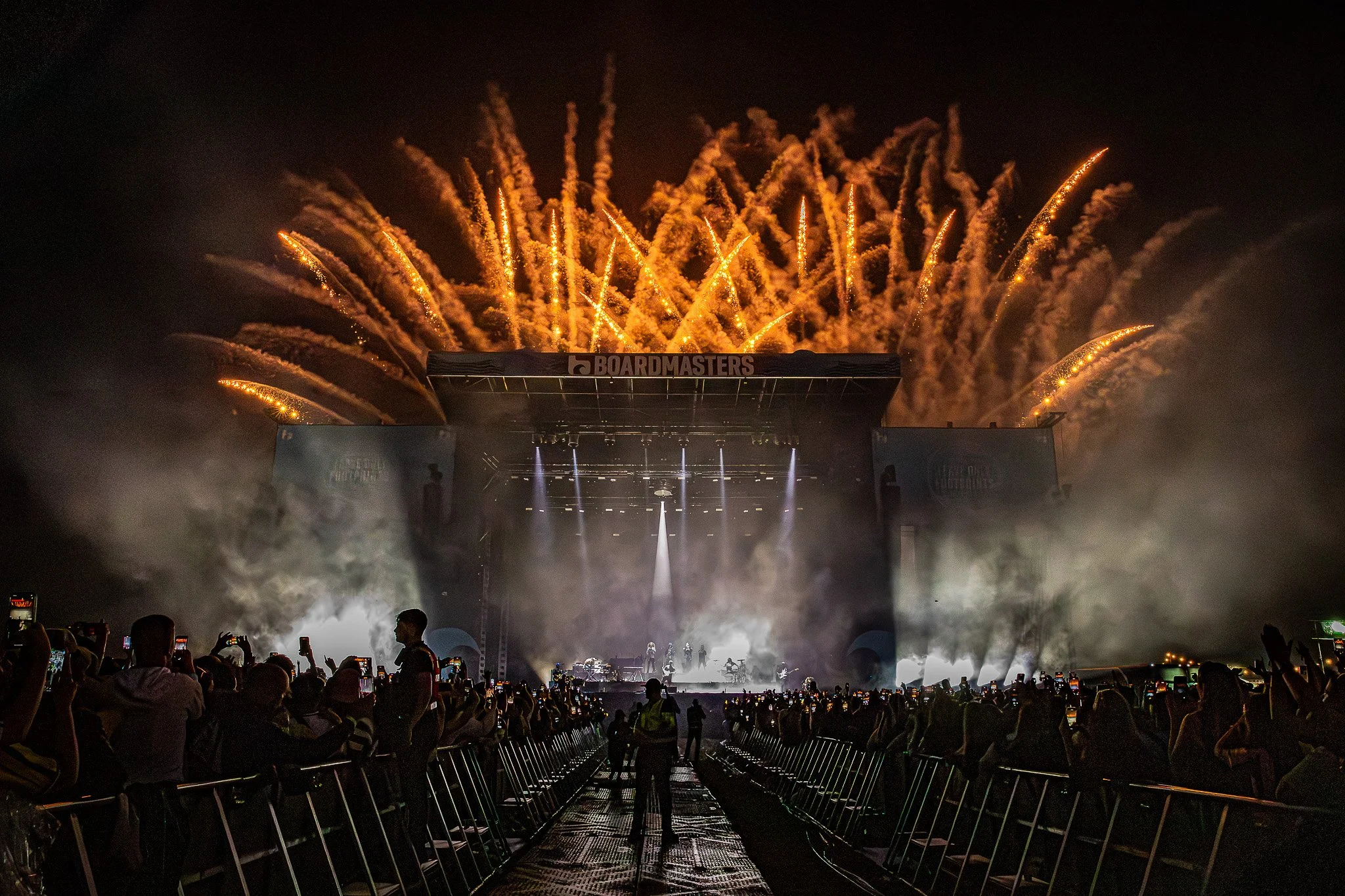 Fireworks display above a stage at an outdoor concert, with audience members taking photos and watching.