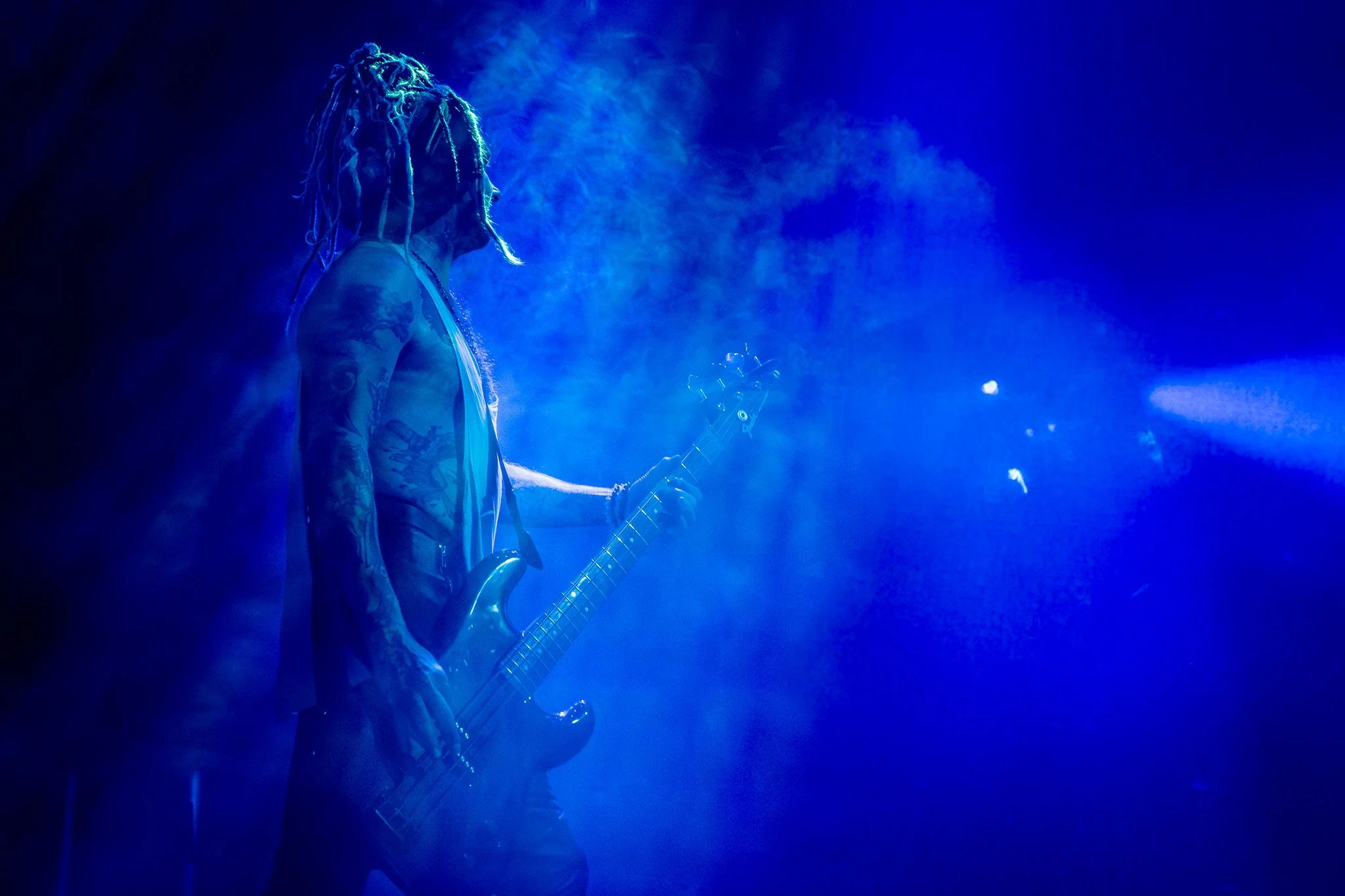 A shirtless man with tattoos and dreadlocks playing a bass guitar on stage illuminated with blue lighting and smoke.