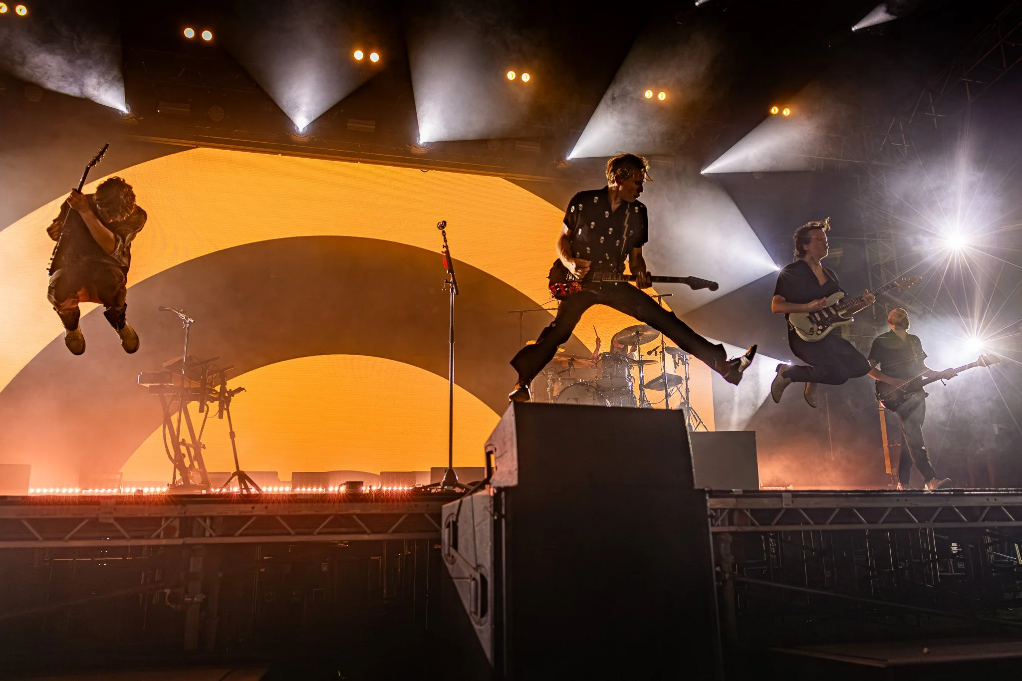 A live band performing on stage with bright stage lights, including three musicians jumping in mid-air, with a large yellow and black backdrop.