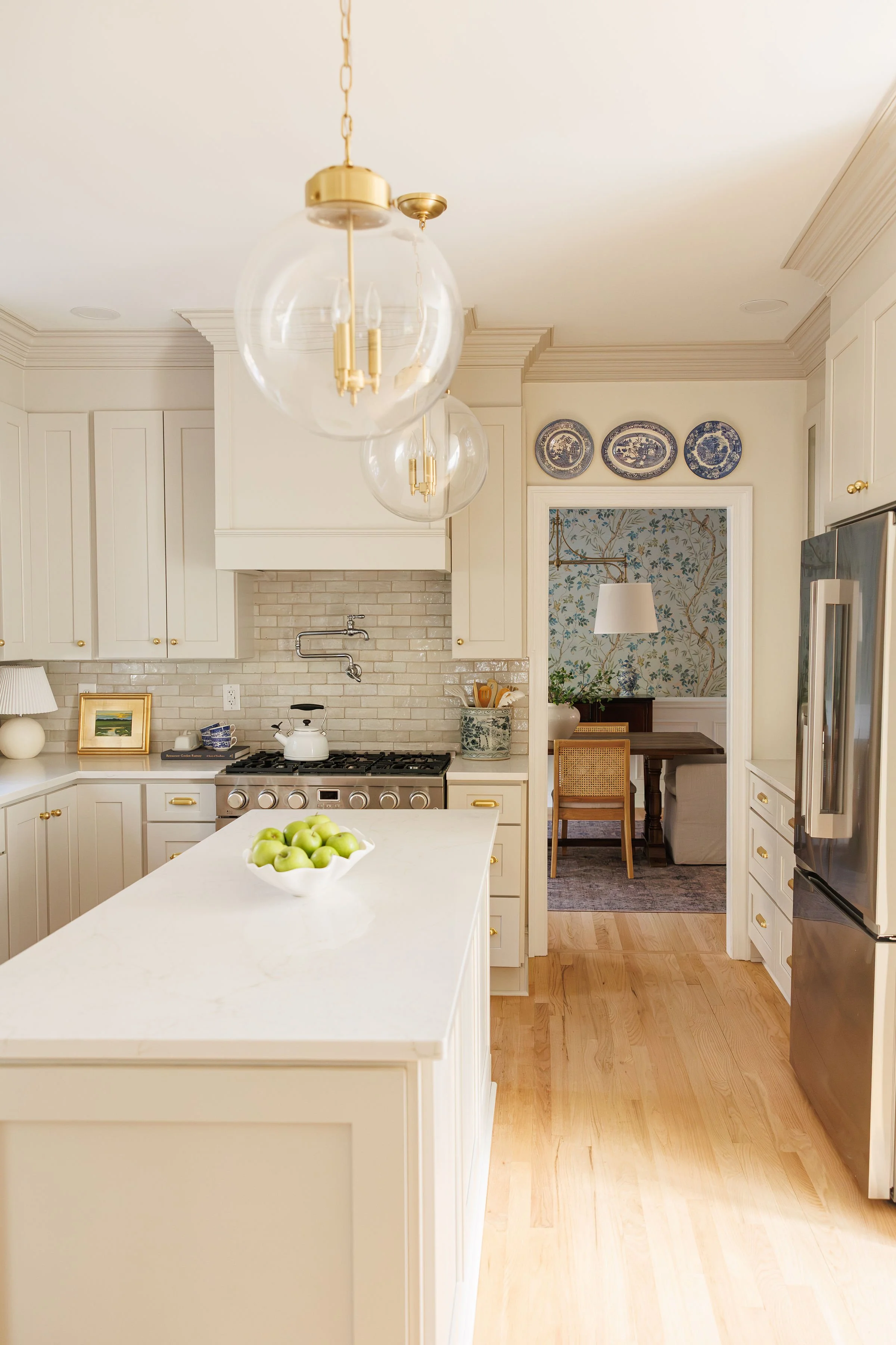 Modern kitchen with white cabinets, a white island with a bowl of green apples, stainless steel refrigerator, wall-mounted oven, and a dining area with wallpapered accent wall and a hanging lamp.