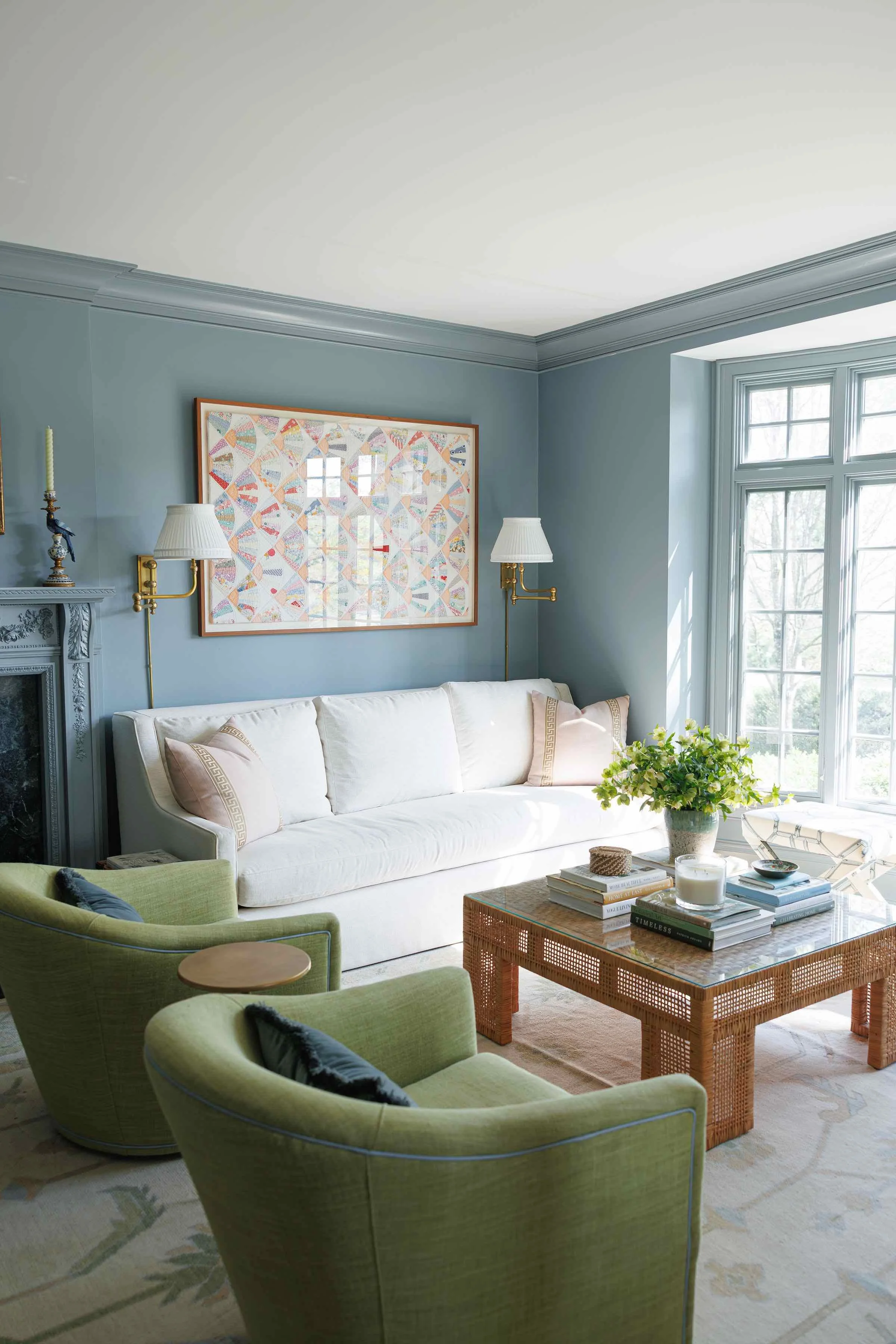 A bright living room with light blue walls, a white sofa, and green chairs. There is a wicker coffee table with books and candles, a window letting in natural light, and decorative wall art.
