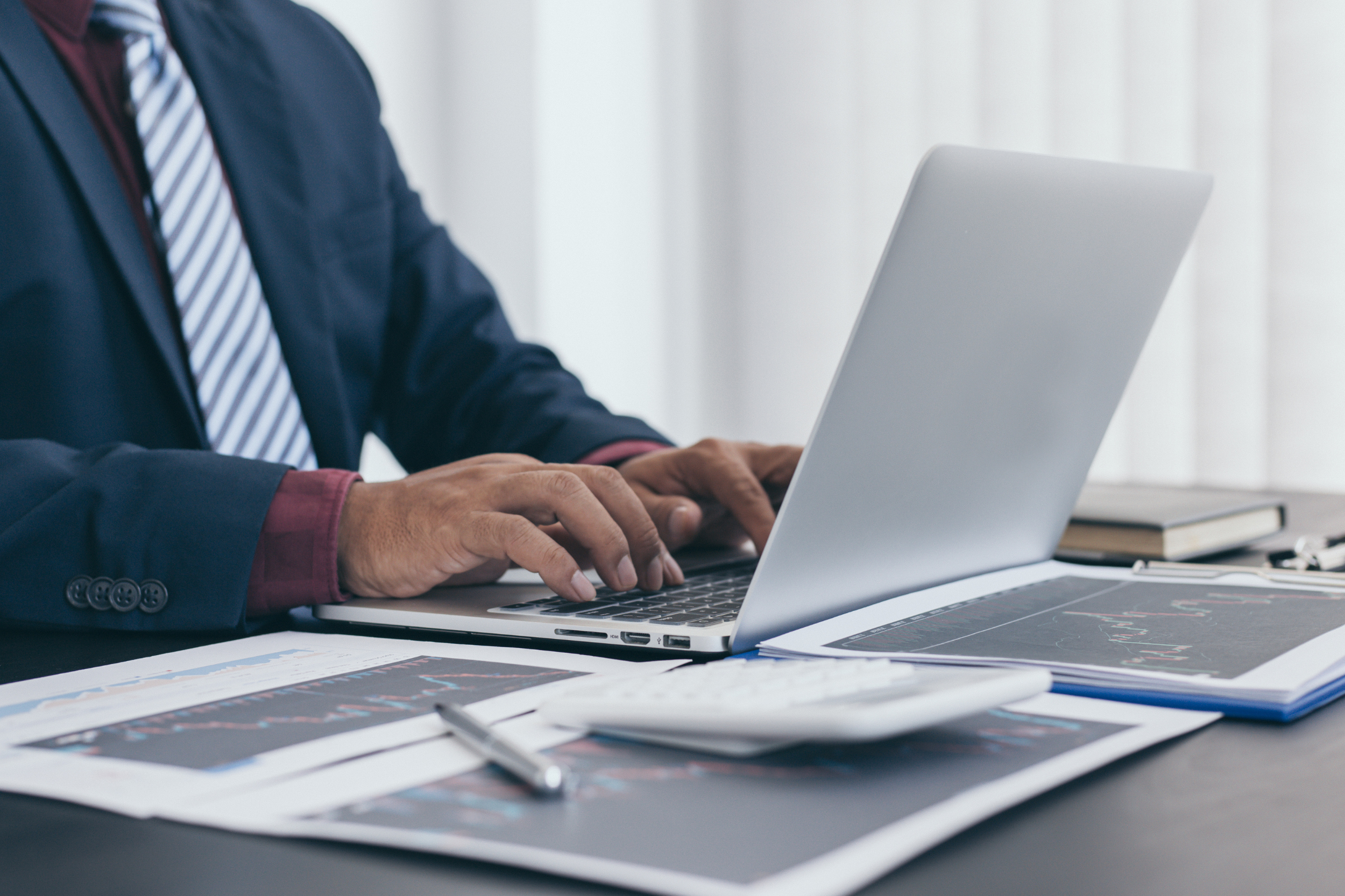 A person in business attire working on a silver laptop in an office setting, with financial charts, documents, a calculator, and a pen on the desk.