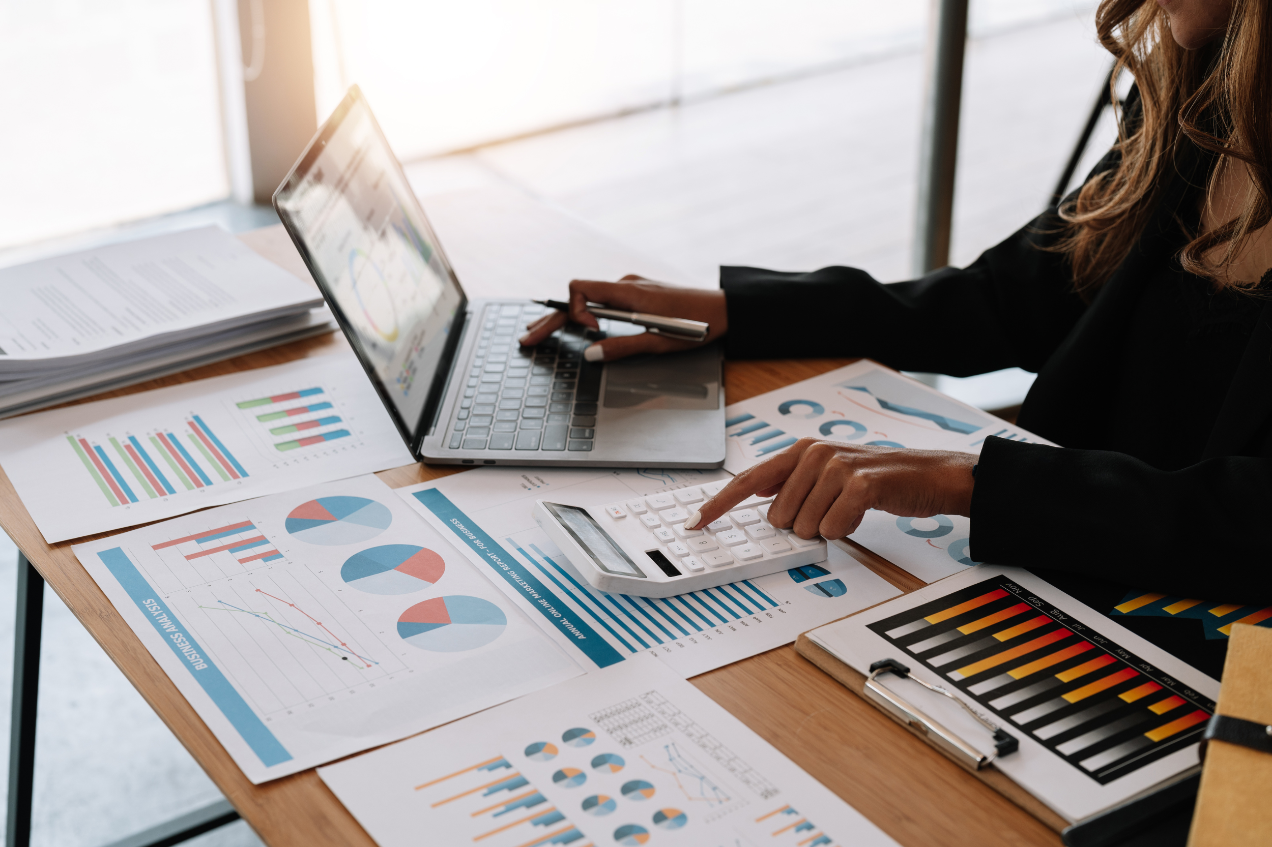 Businesswoman analyzing financial data on printed charts and graphs, using a calculator and a laptop on a wooden table.