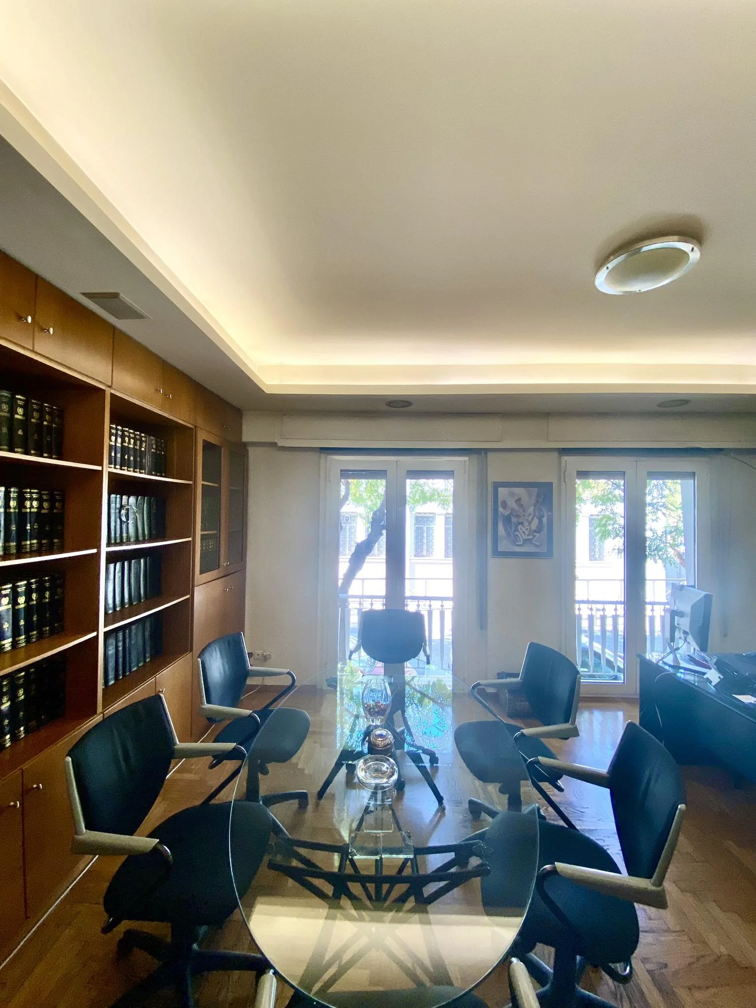 An office conference room with six black chairs around a glass table, a wooden bookshelf on the left, and two large windows in the background.