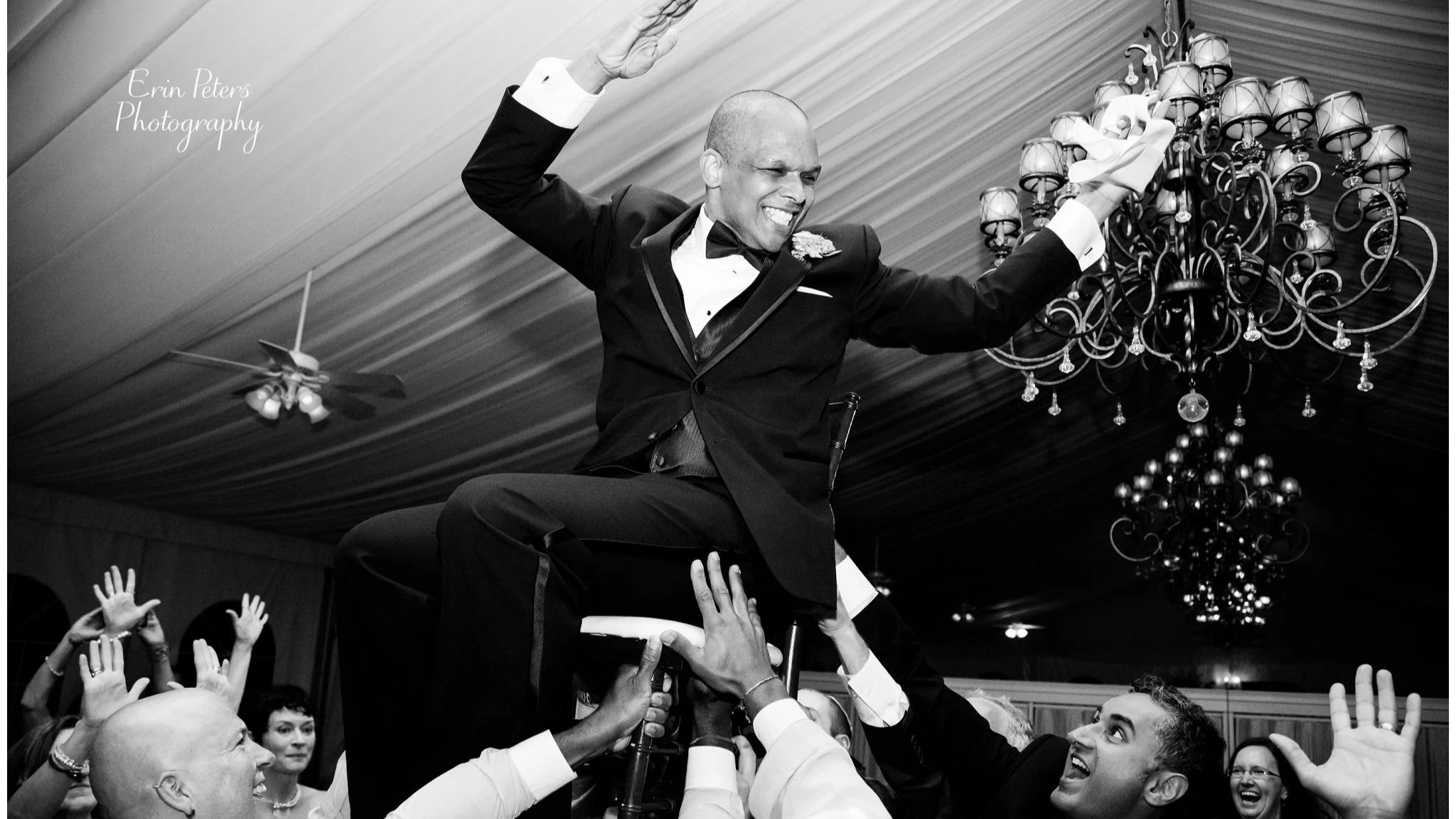 The groom is being lifted on a chair during a wedding celebration, smiling and raising his hands, surrounded by guests and family at West Hills Country Club