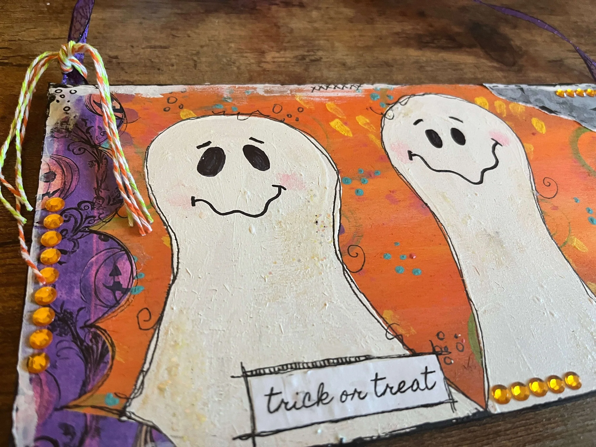Handmade Halloween decoration featuring two smiling ghost drawings on a colorful background with purple and orange accents, decorated with orange rhinestones, and a sign that says 'trick or treat.'