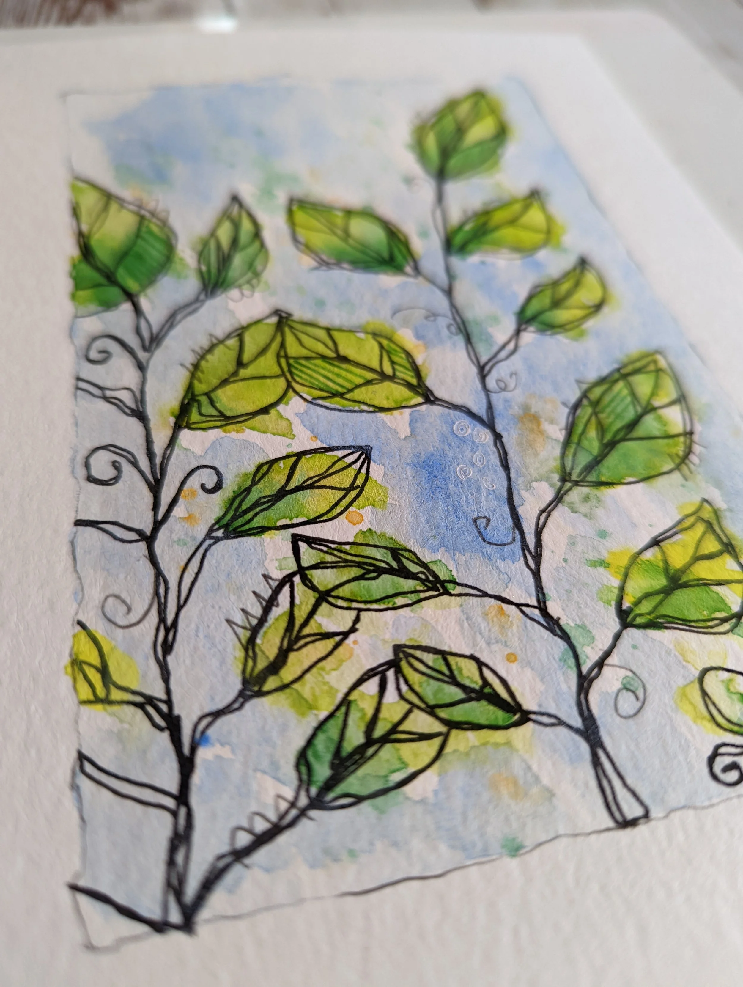 A watercolor painting of green leaves on black inked branches with a light blue and yellow background.