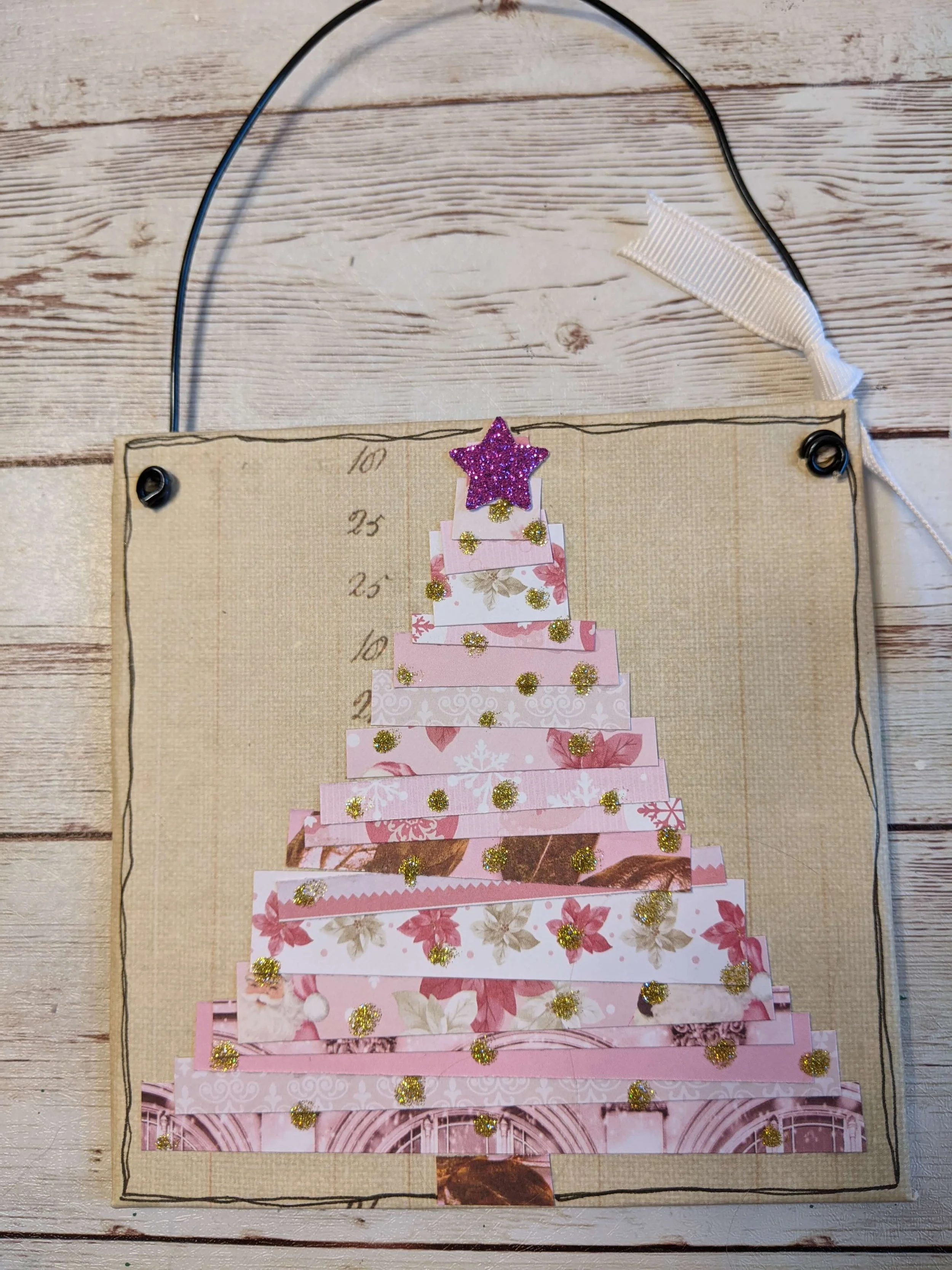 A handmade Christmas decoration featuring a collage of pink and floral patterned paper strips arranged in the shape of a Christmas tree, decorated with gold glitter dots and topped with a purple glitter star. The decoration is outlined with a black line and has a white ribbon on the top right corner for hanging.