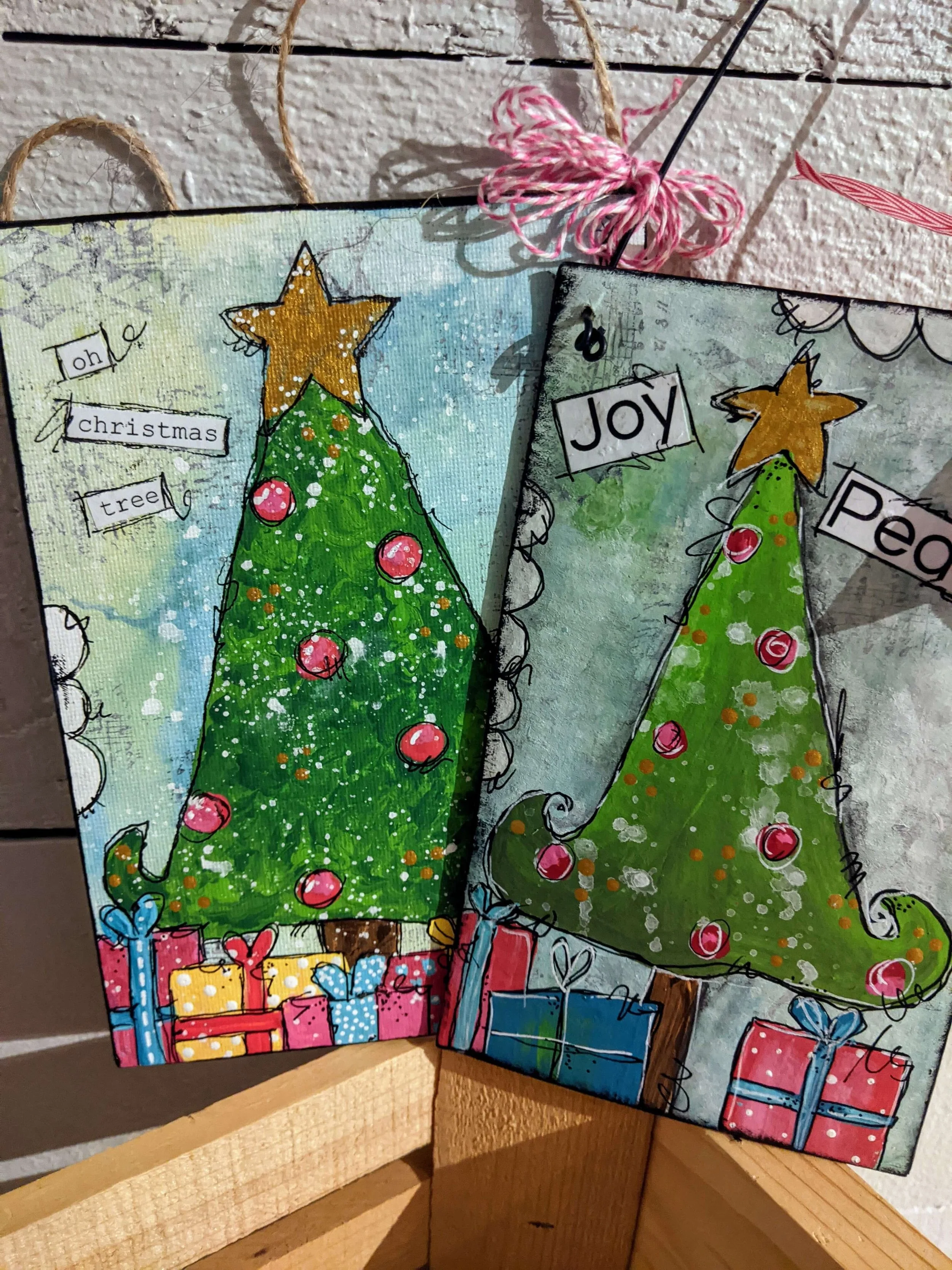 Two colorful Christmas cards featuring decorated Christmas trees with ornaments and gifts underneath. The cards are painted with a whimsical style and have handwritten words such as 'Oh Christmas Tree', 'Joy', and 'Peace' attached with small black clips.