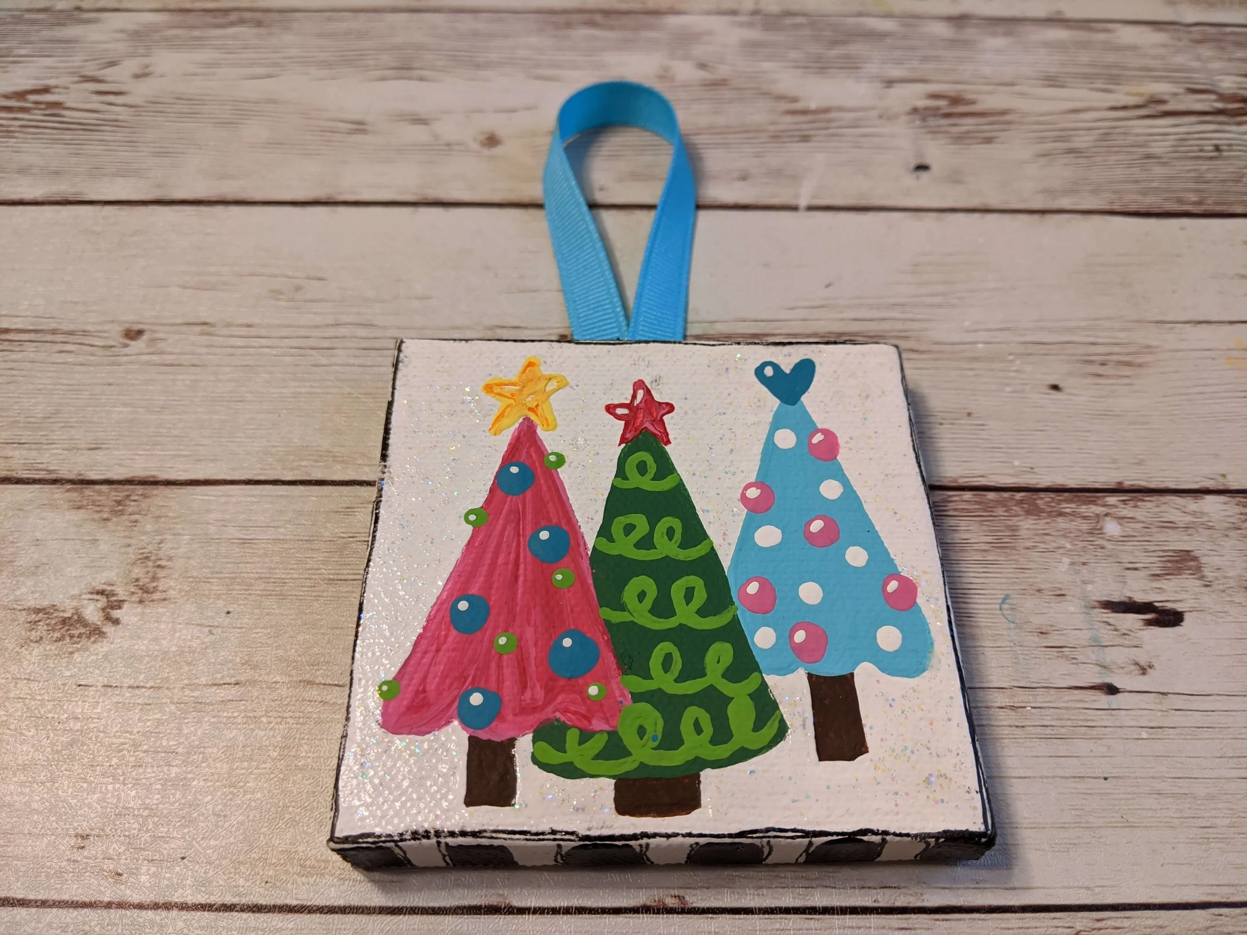 Decorated Christmas hand made ornament with painted Christmas trees and a blue ribbon handle on a wooden surface.