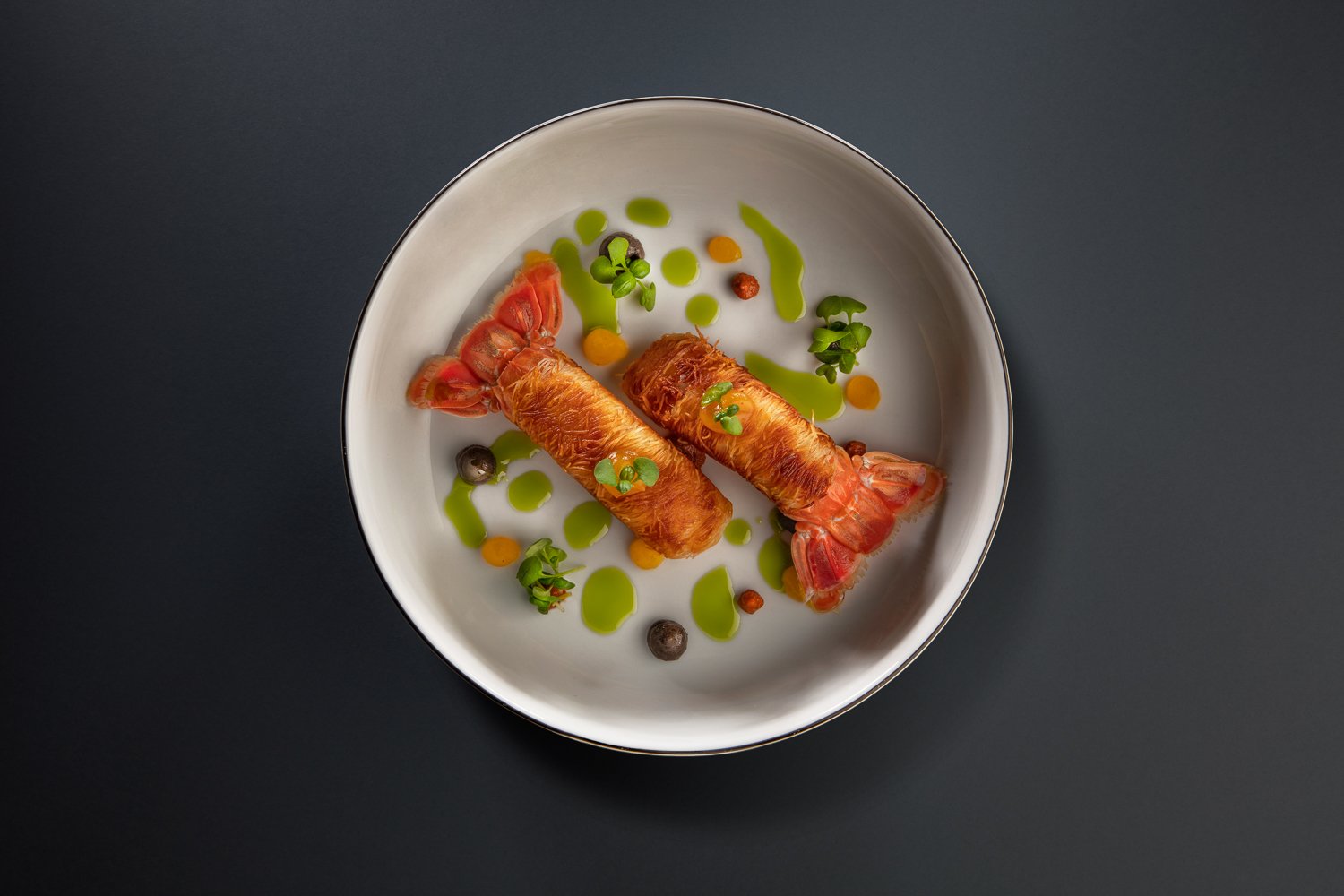 Gourmet dish with two crispy rolls filled with green vegetables and colored sauce, decorated with small portions of green and orange sauce.
