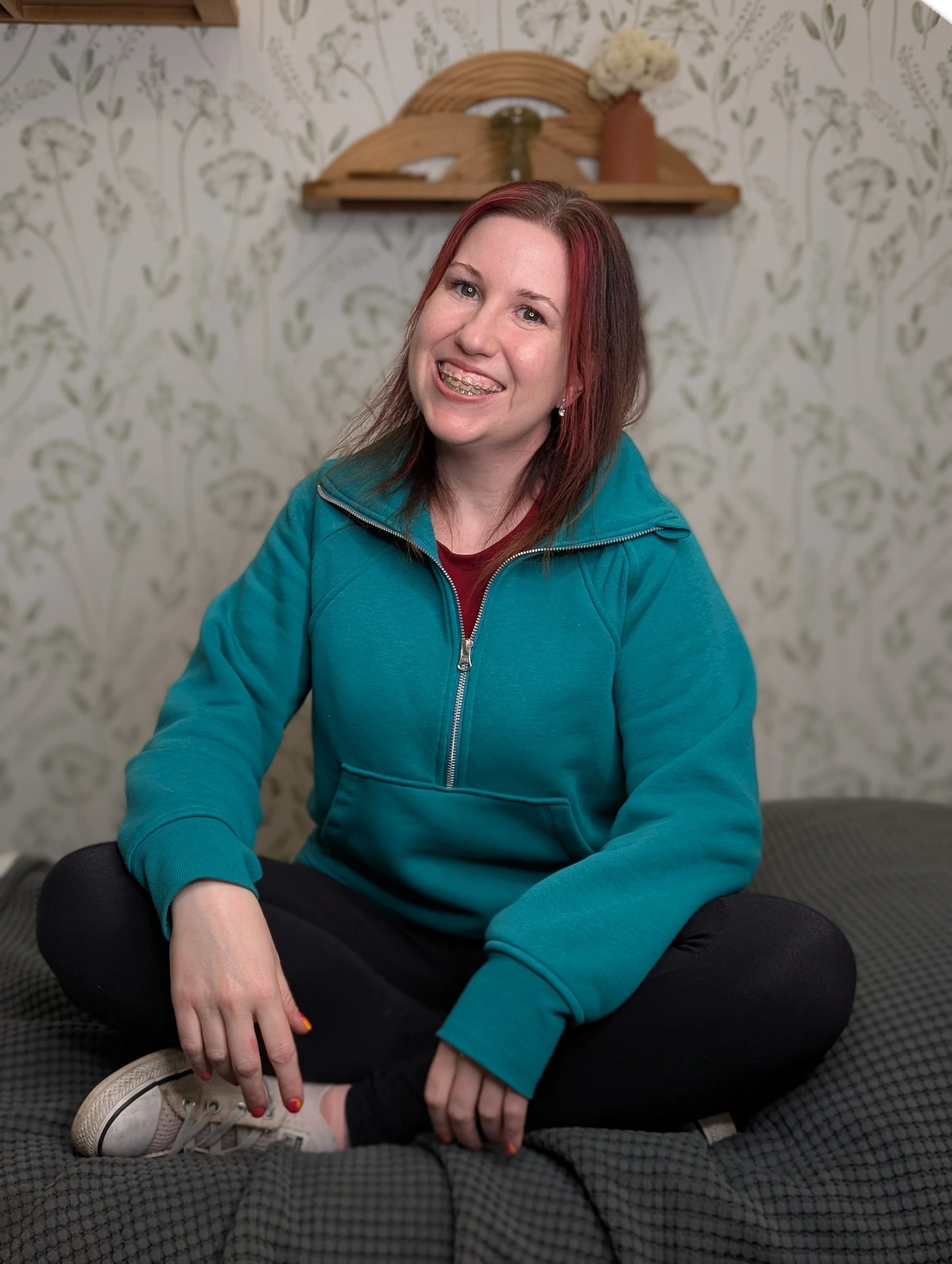 A woman with red hair and braces sitting cross-legged on a massage table, wearing a sweater and black pants, smiling at the camera. There is a floral wallpaper behind her and a small wooden shelf with decorative vases and a flower on top.