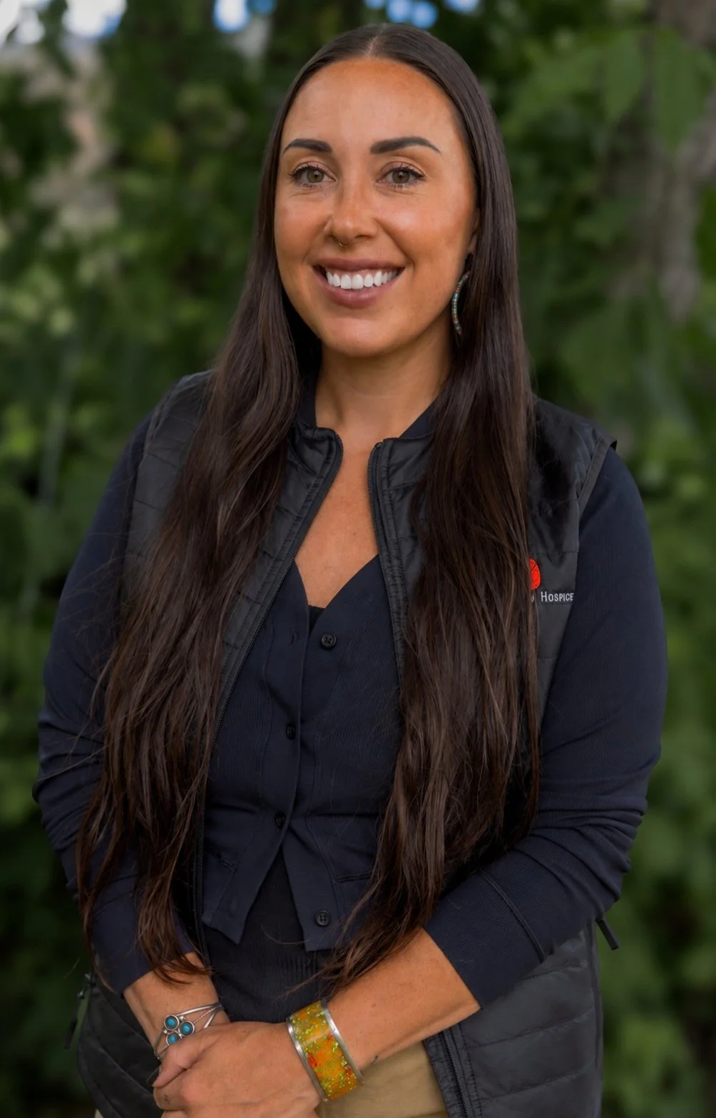 Portrait of a smiling woman with long dark hair, wearing a gray vest with a logo, a black shirt, and colorful feather necklace, standing in front of a turquoise wooden background.