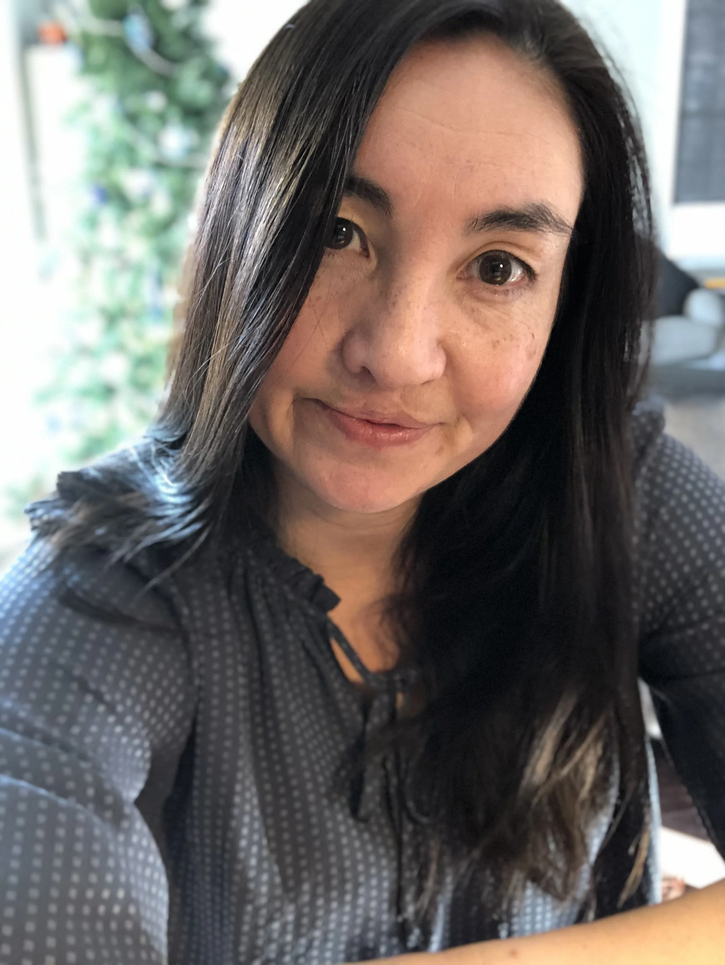 A woman with long black hair, wearing a black and gray polka dot shirt, looking at the camera with a slight smile, background of a well-lit indoor space with blurred plants and furniture.
