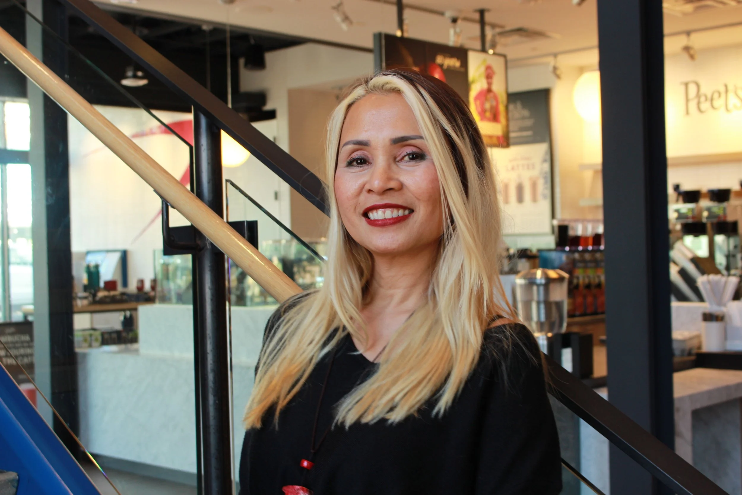 A woman with blonde hair wearing a black top and red lipstick standing inside a coffee shop, smiling at the camera near a staircase.