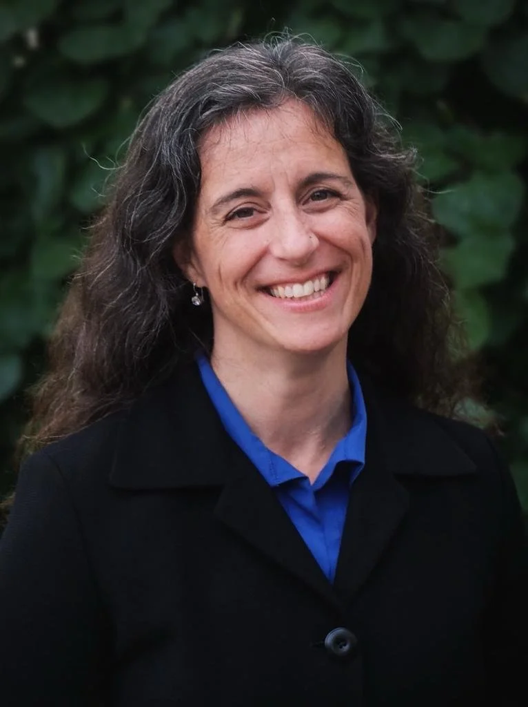 A woman with long, wavy gray hair smiling in front of green foliage, wearing a black blazer and a blue shirt.