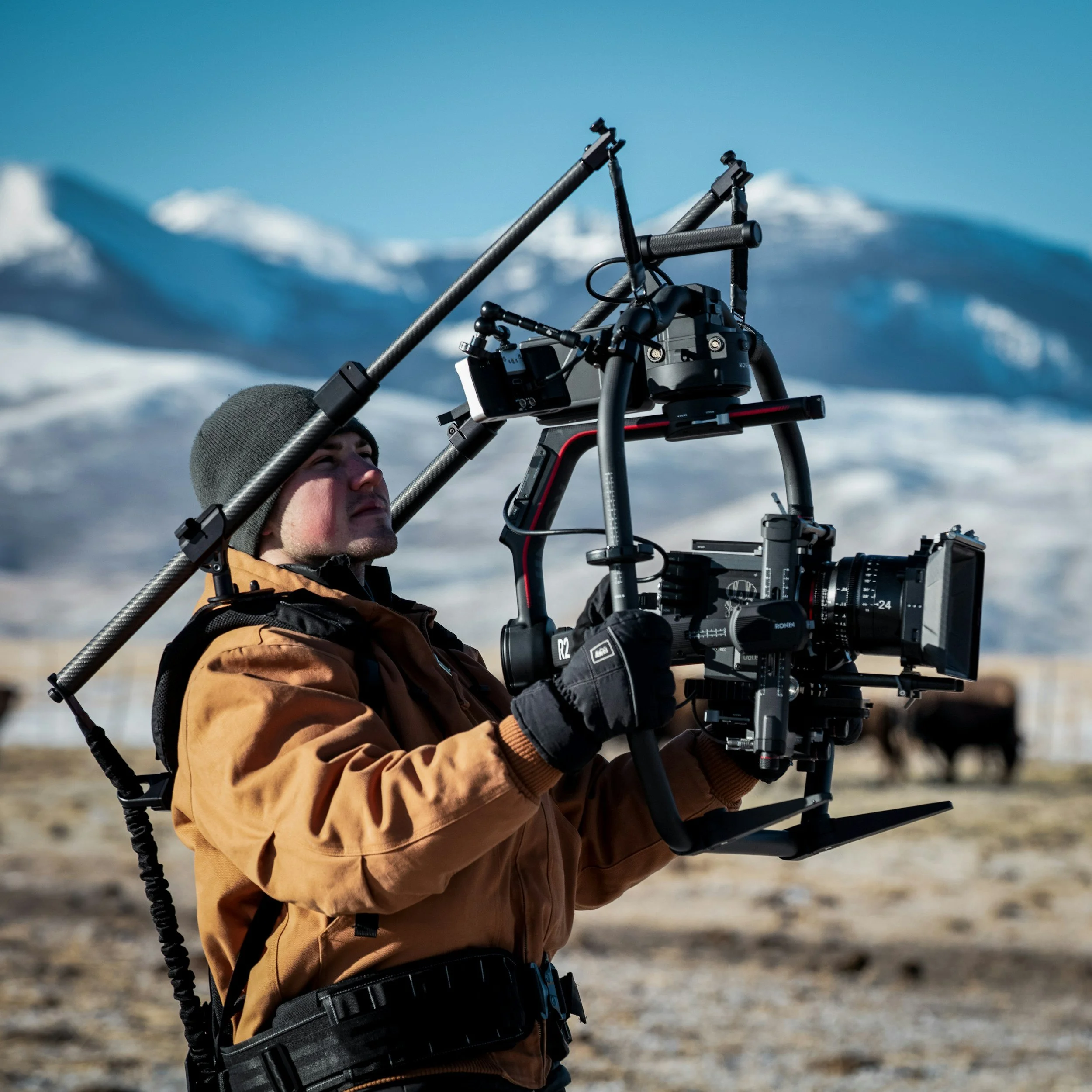 A man in winter clothing operating a professional camera rig outdoors with snow-covered mountains in the background.