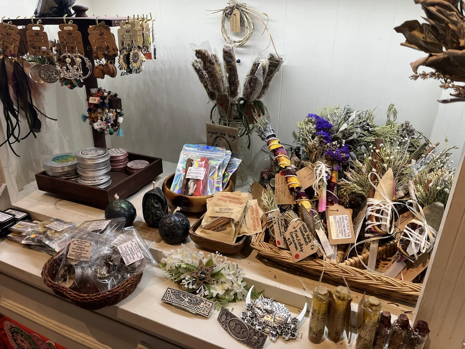 A display of various small decorative items, jewelry, dried flowers, and herbal products in a shop. Items include earrings, herbal bundles, candles, and herbal salves.