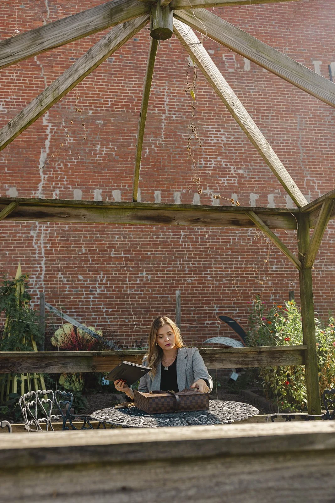 Kerri M. Roberts sitting at a garden table with a brick wall background, looking into a bag and holding a book or folder.