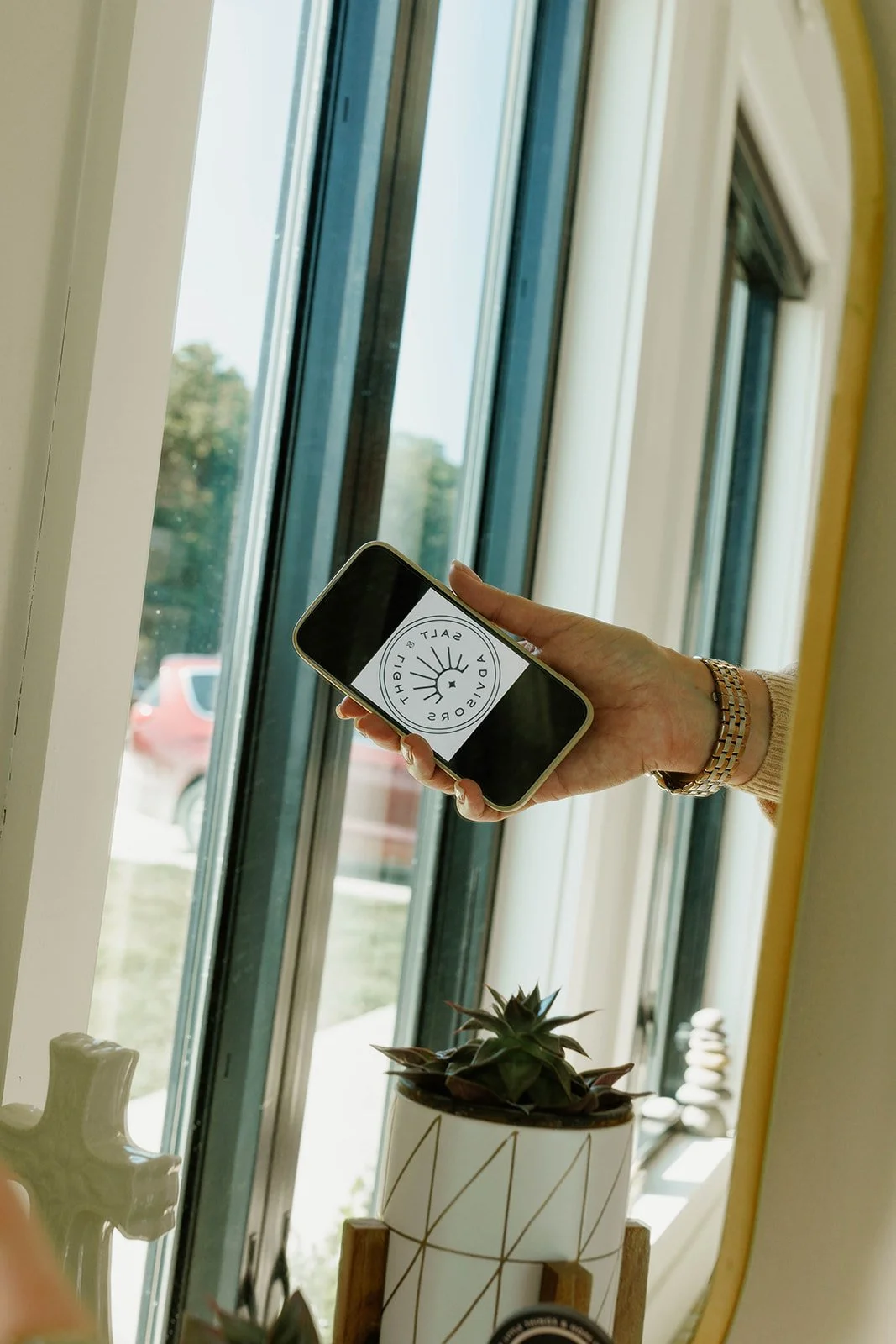 Person holding a smartphone with a logo that says 'Salt & Light Advisors' in front of a large window with trees outside and a potted plant nearby.