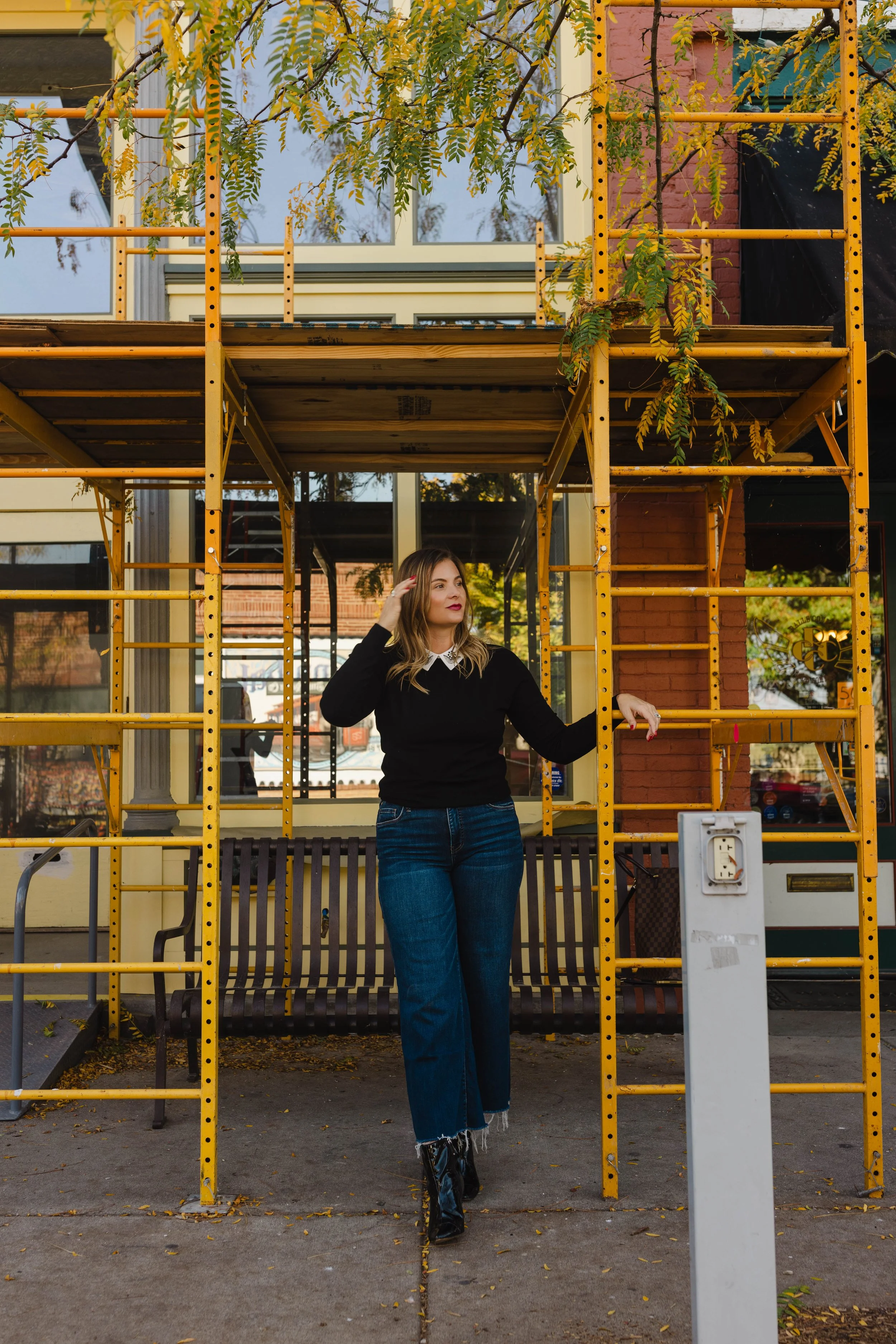 A woman with long blonde hair wearing a black sweater, white collared shirt, blue jeans, and black boots stands in front of a yellow scaffolding structure outside a storefront with large windows, some trees with yellow leaves are visible.