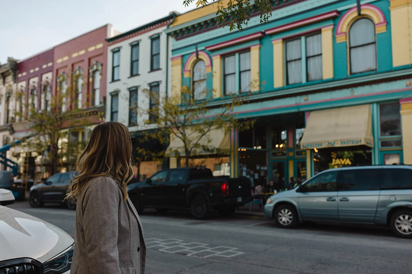 Kerri M. Roberts standing on the sidewalk looking towards colorful, decorated shops on a city street. Several parked cars are visible, and the sky indicates it is late afternoon.