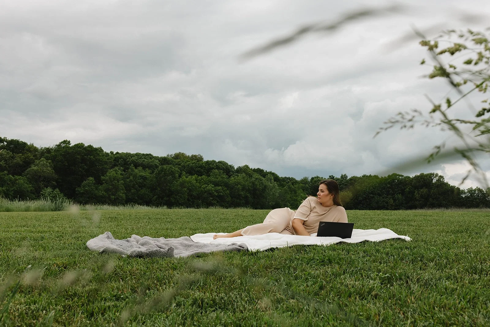 Kerri M. Roberts lying on a white blanket on a grassy field under a cloudy sky, with a laptop nearby.