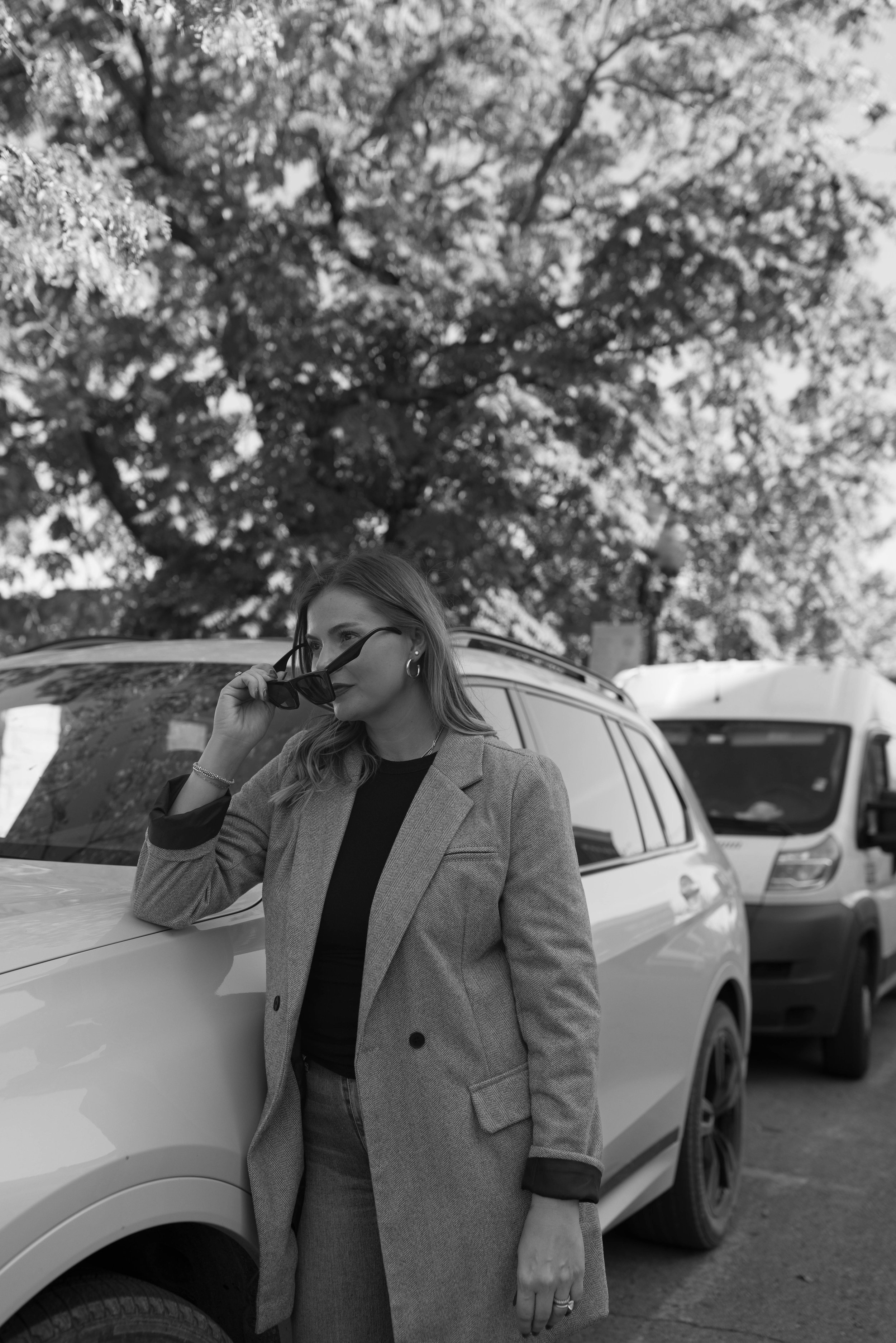 A woman with long hair wearing a blazer and earrings stands next to a car, holding sunglasses near her face, with trees and parked vehicles in the background.