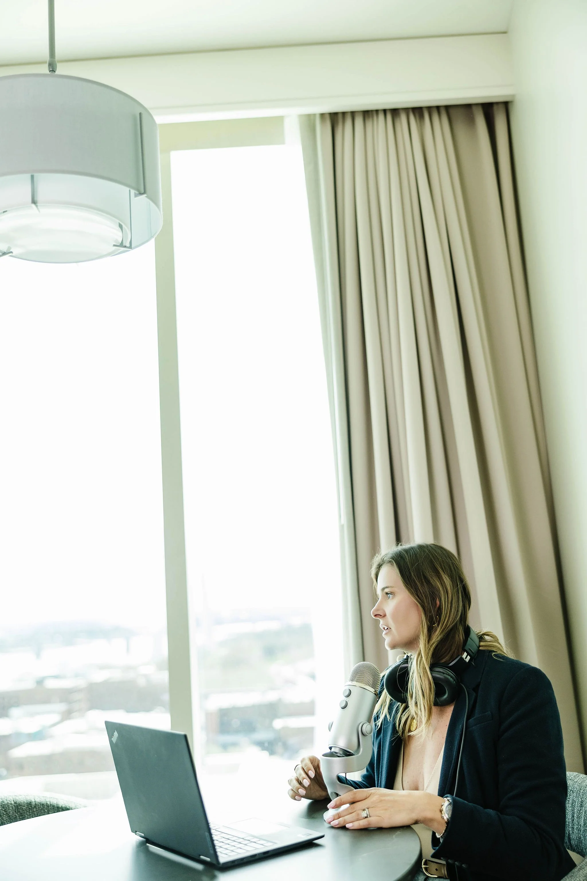 Kerri M. Roberts sitting at a table with a laptop, wearing headphones around her neck, speaking into a microphone in a bright room with large windows and curtains.