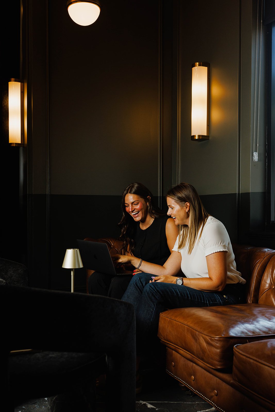 Kerri M. Roberts and an employee sitting on a leather sofa in a dimly lit room, looking at a laptop, smiling and having a conversation.