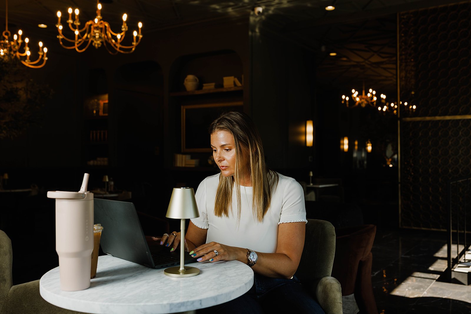 Kerri M. Roberts working on her laptop at a small round marble table in a dimly lit, elegant restaurant or lounge with chandeliers and warm lighting.