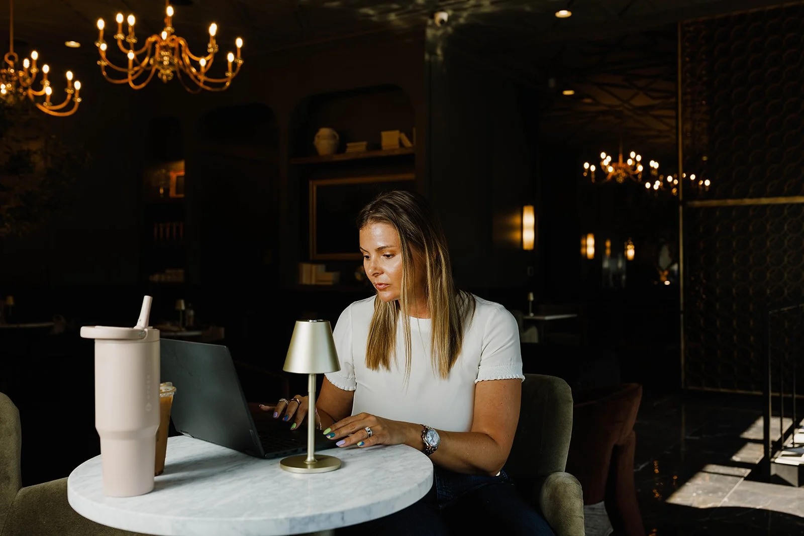 Kerri M. Roberts working on her laptop at a small round marble table in a dimly lit, elegant restaurant or lounge with chandeliers and warm lighting.