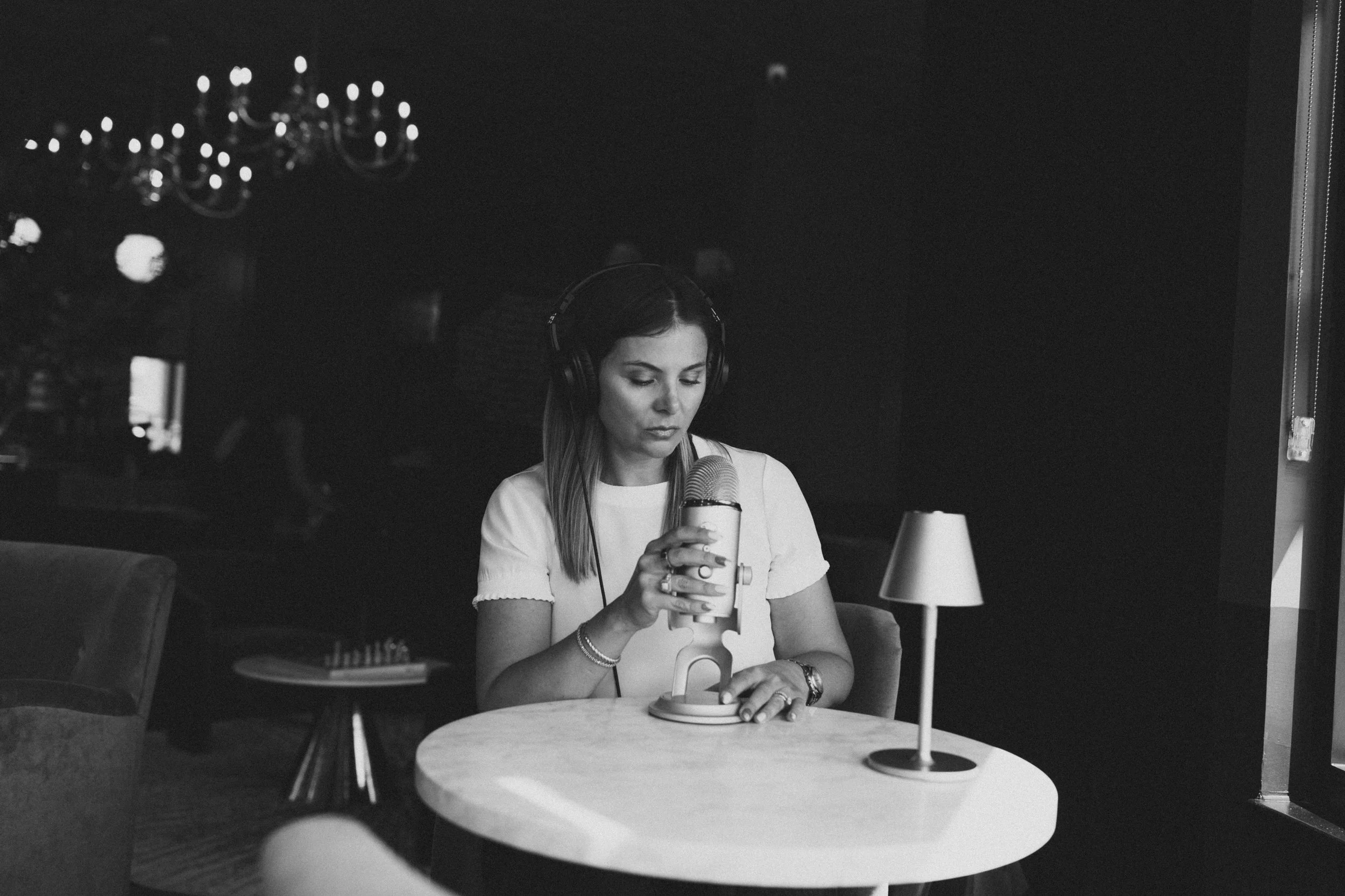 Woman with headphones speaking into a microphone at a round table in a dimly lit room.