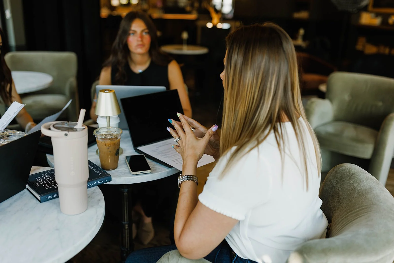 Kerri M. Roberts is sitting at a table in a cafe, holding her hands together while speaking. There are two other women visible in the background, one with dark hair and another mostly out of frame.