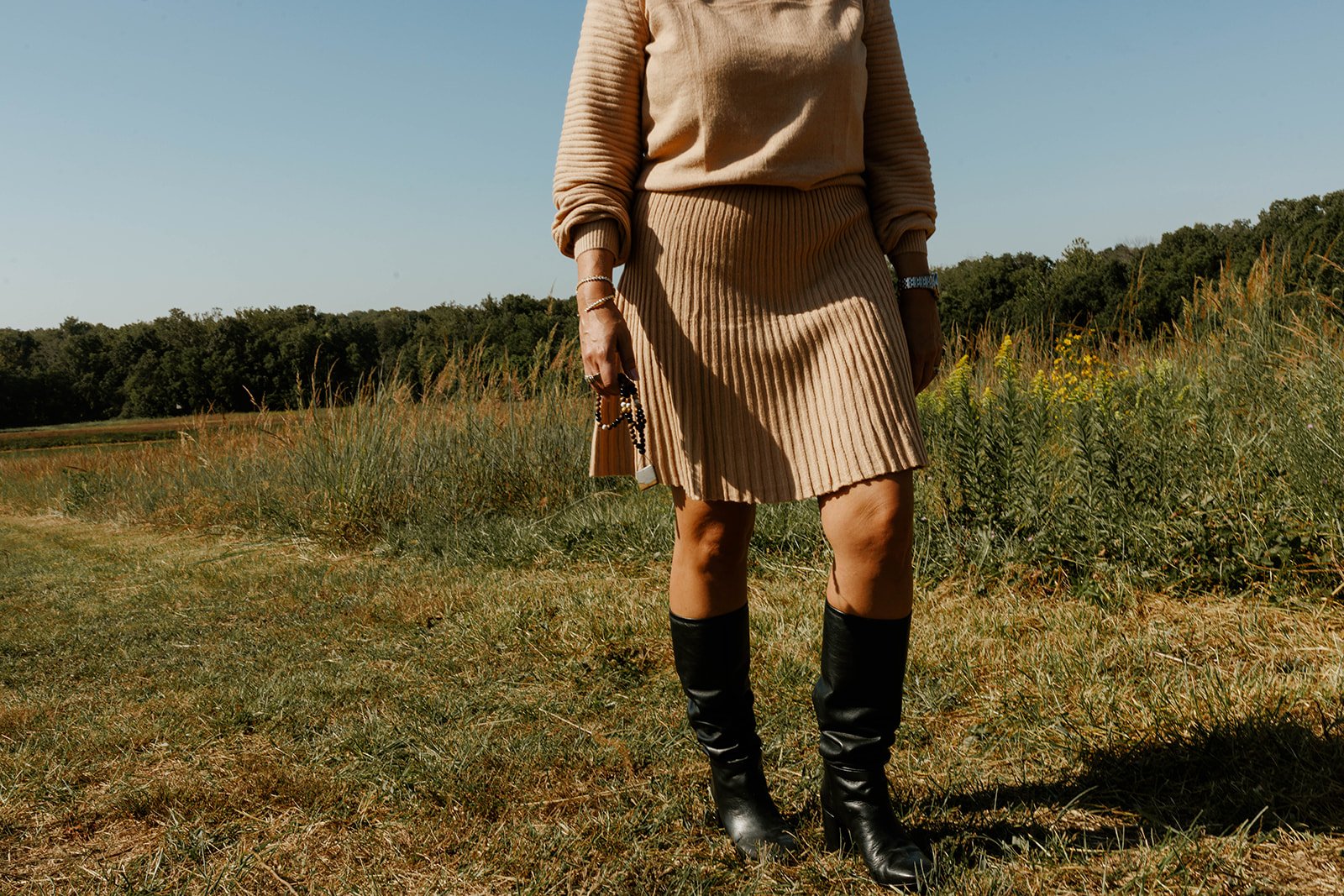 Kerri M. Roberts standing outdoors in a field of grass and wildflowers, wearing a beige sweater, a beige pleated skirt, black knee-high boots, and holding a small clutch purse with beaded accessories.