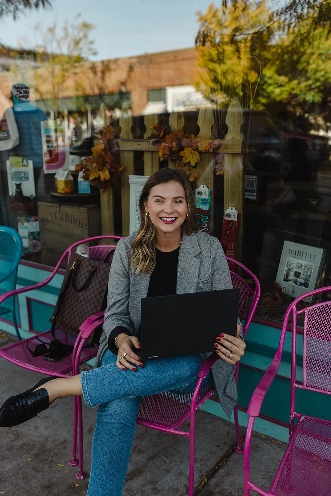 Kerri M. Roberts sitting on a pink metal bench outside a storefront, smiling, holding a black folder, with a brown Louis Vuitton bag beside her. She is wearing a gray blazer, black top, and blue jeans, with red painted nails.