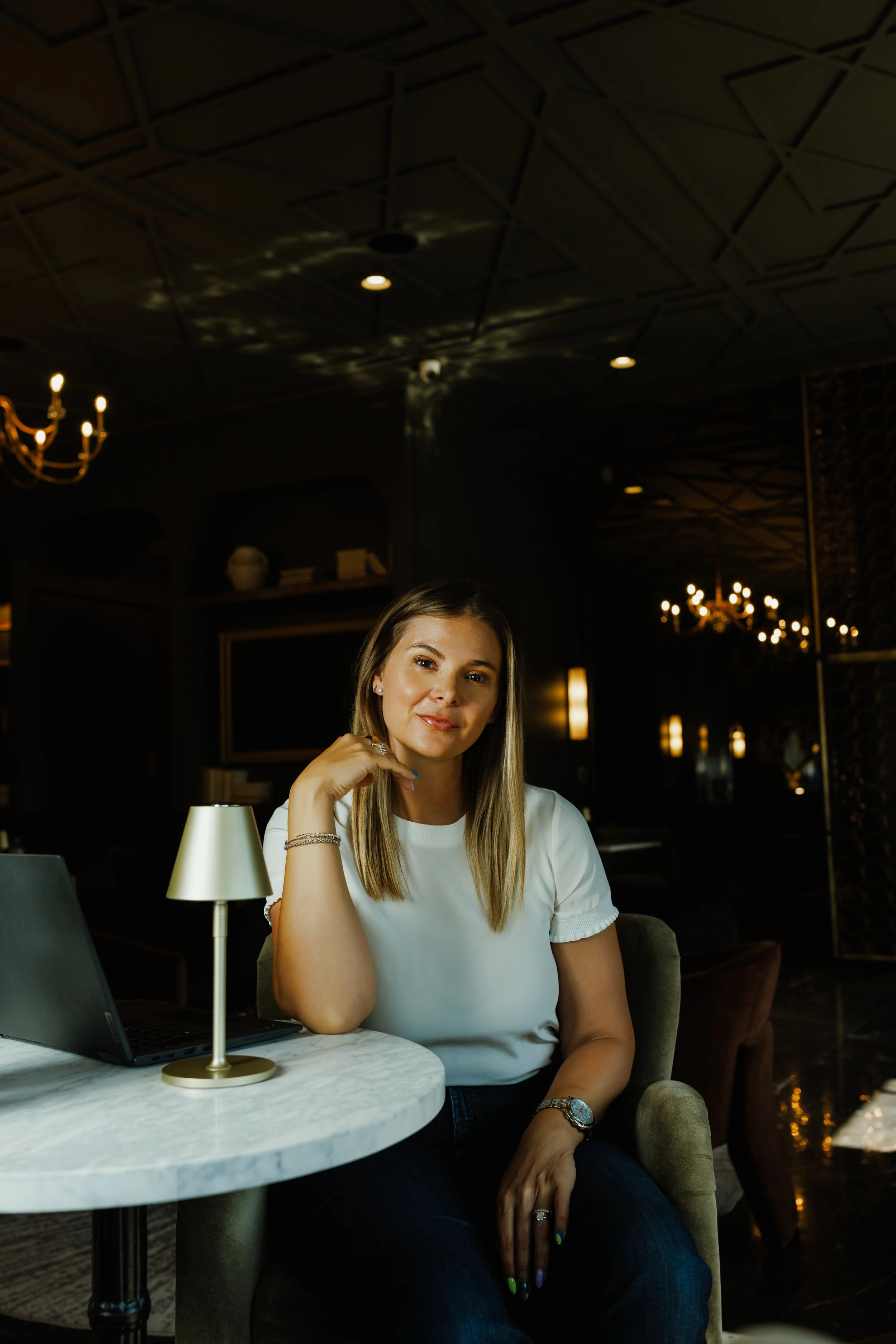 A young woman with shoulder-length blonde hair, sitting at a marble table in a dimly lit, elegant lounge or hotel lobby, with a laptop open in front of her. She is wearing a white blouse, jewelry, and has her right hand resting near her face, smiling softly.