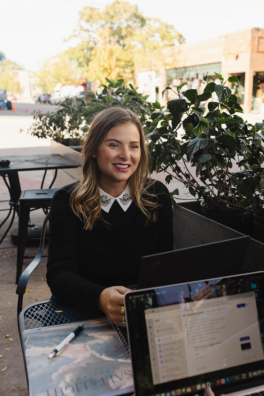 Kerri M. Roberts smiling while looking at her laptop outdoors, with potted plants and a street scene in the background.