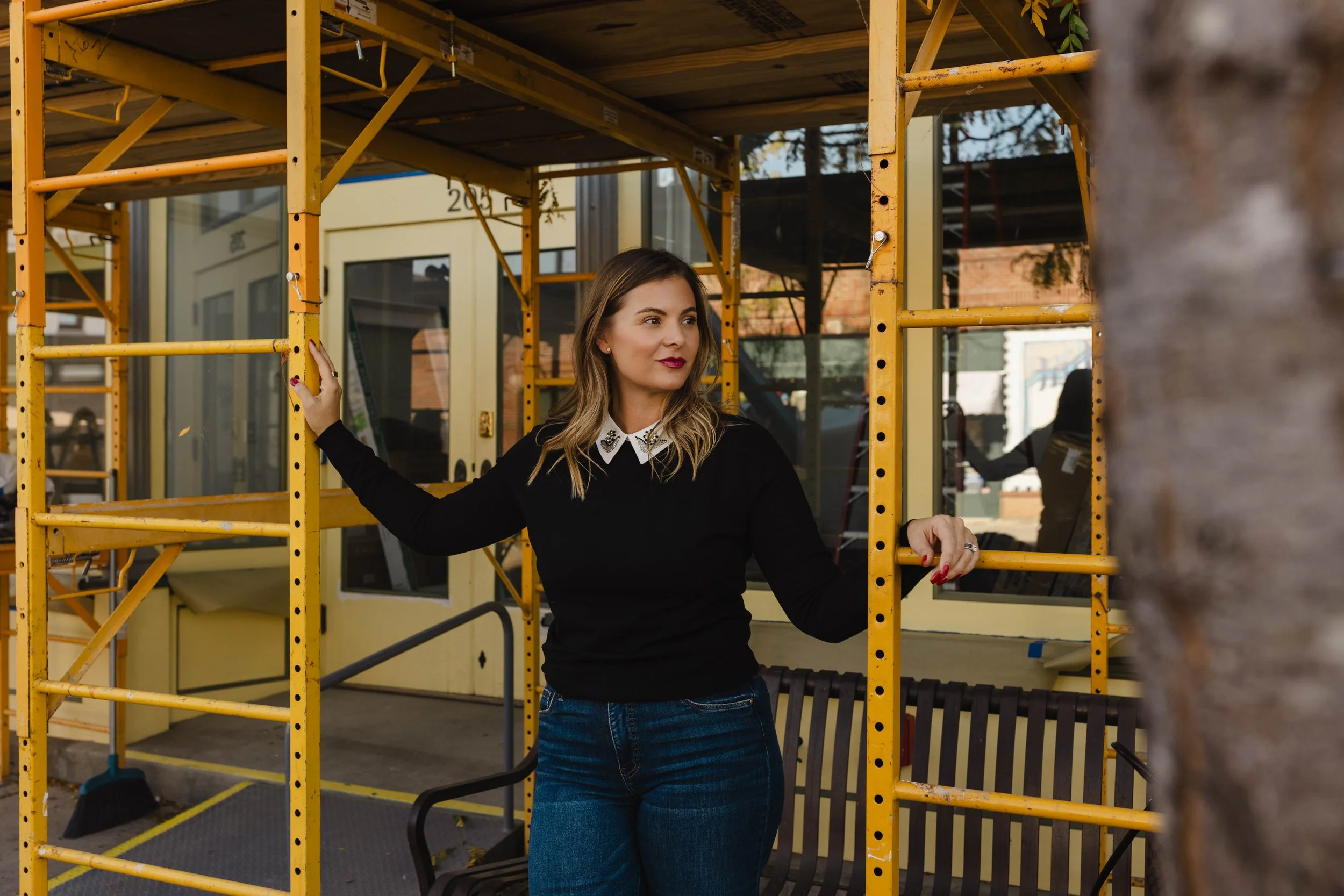 A woman standing amid yellow scaffolding outside a building, looking to the side with her left hand on the scaffolding and right hand on another part, wearing a black sweater with a white collar, blue jeans, and red lipstick.