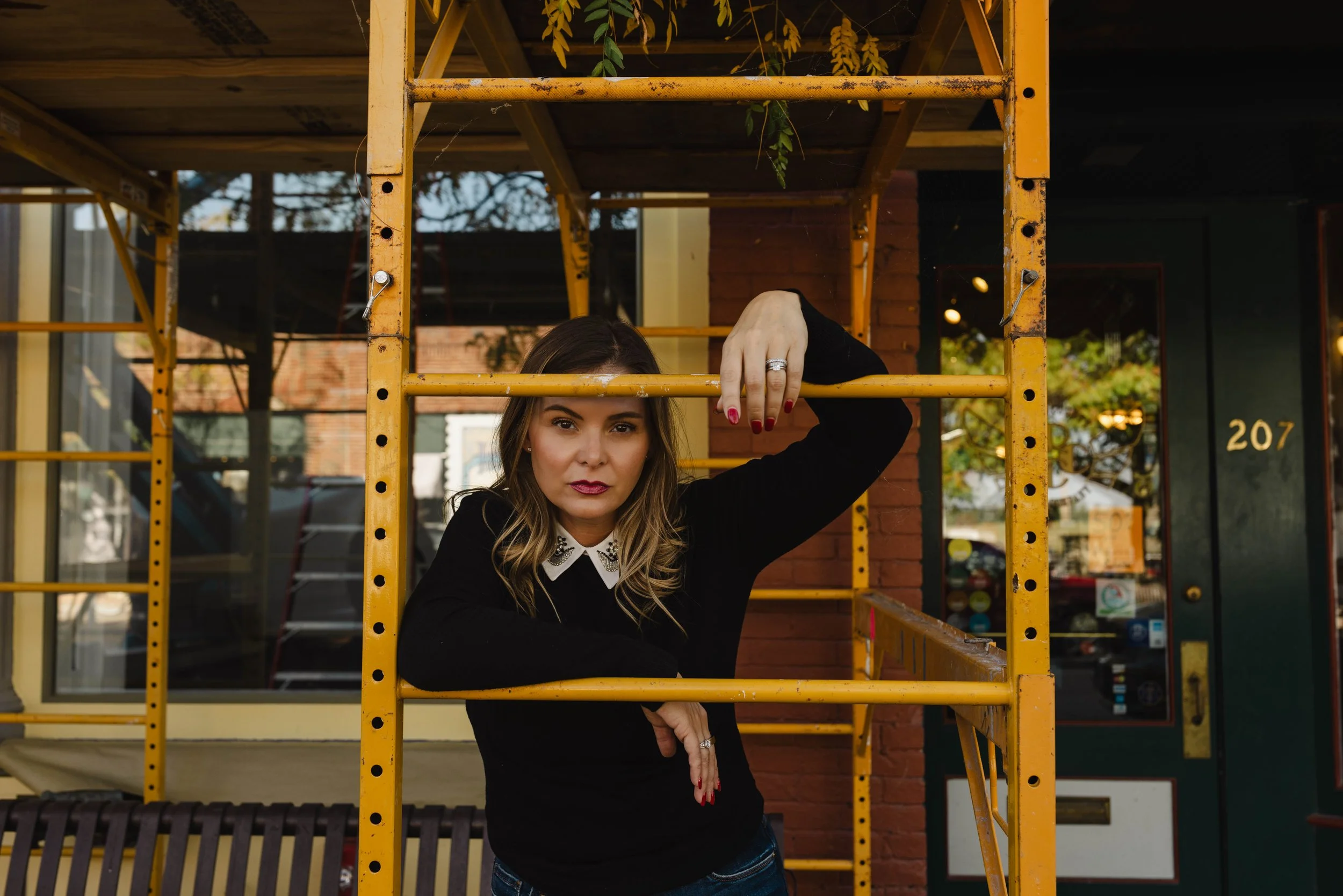 A woman with wavy hair, wearing a black sweater with a white collar, poses with her arm resting on a yellow metal scaffolding outside a building with a green door numbered 207.