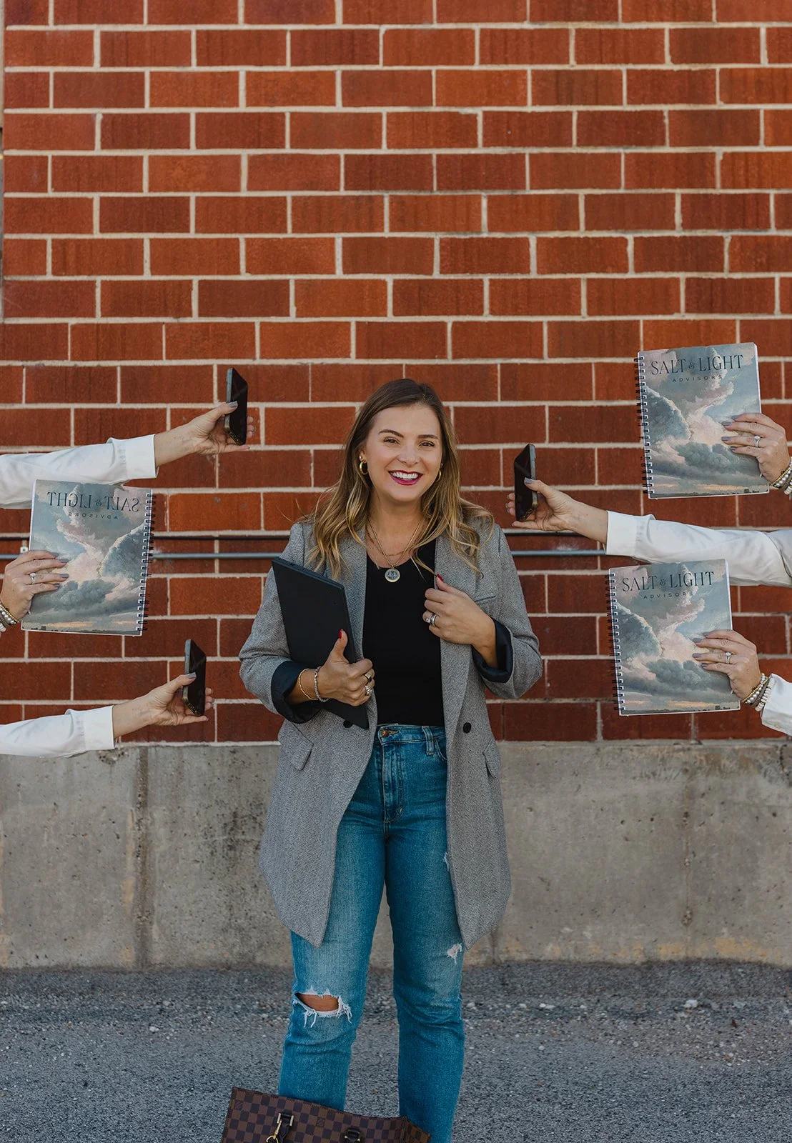 Kerri M. Roberts standing in front of a brick wall, smiling, holding a folder, surrounded by six hands holding smartphones and six books titled 'Salt & Light Advisors'.