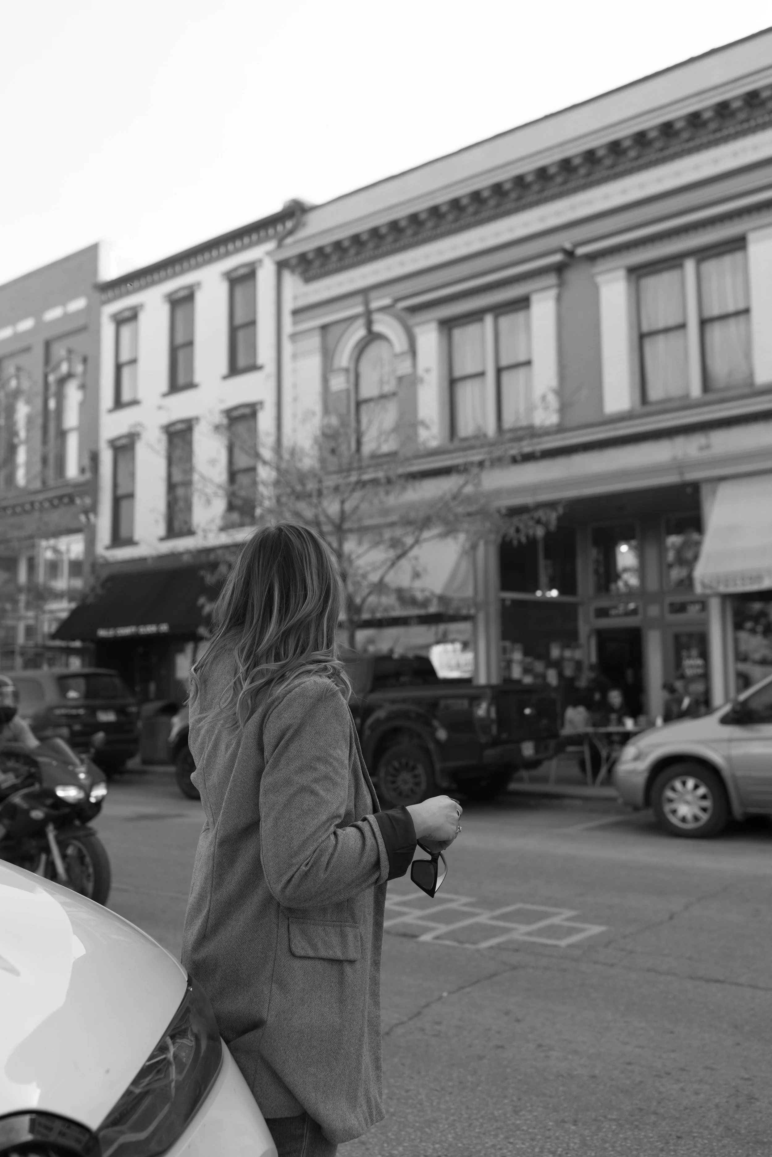A woman in a coat holding sunglasses, standing on a city street with parked cars and historic buildings in the background.