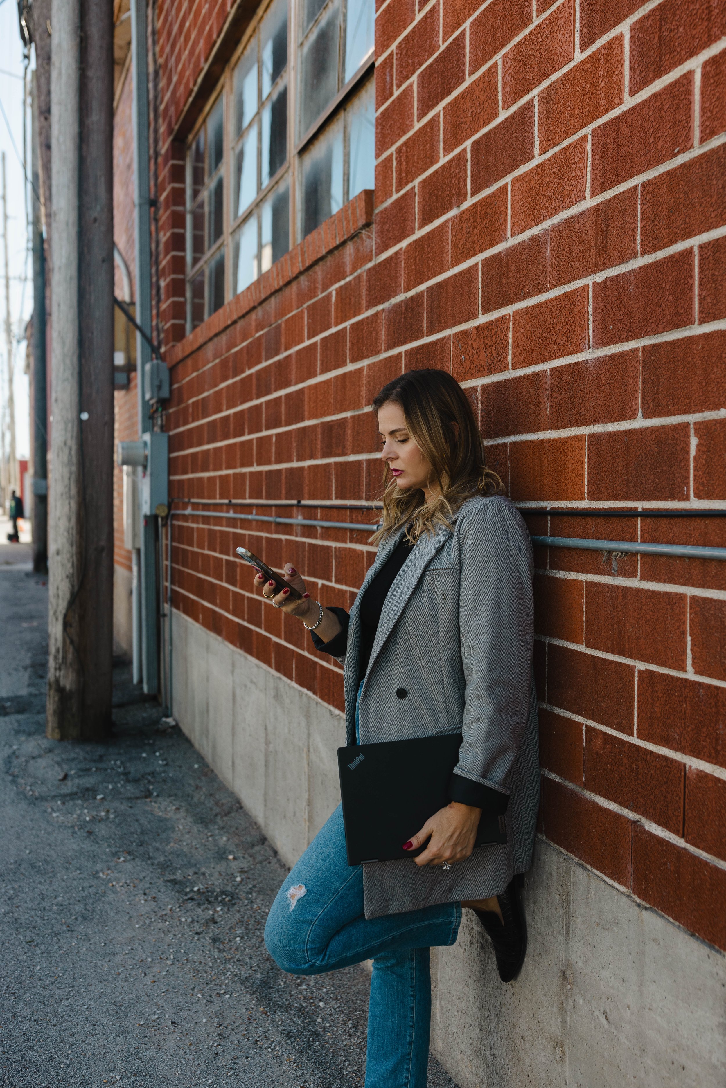 A woman leaning against a brick wall while looking at her phone, holding a black laptop, with a street scene in the background.