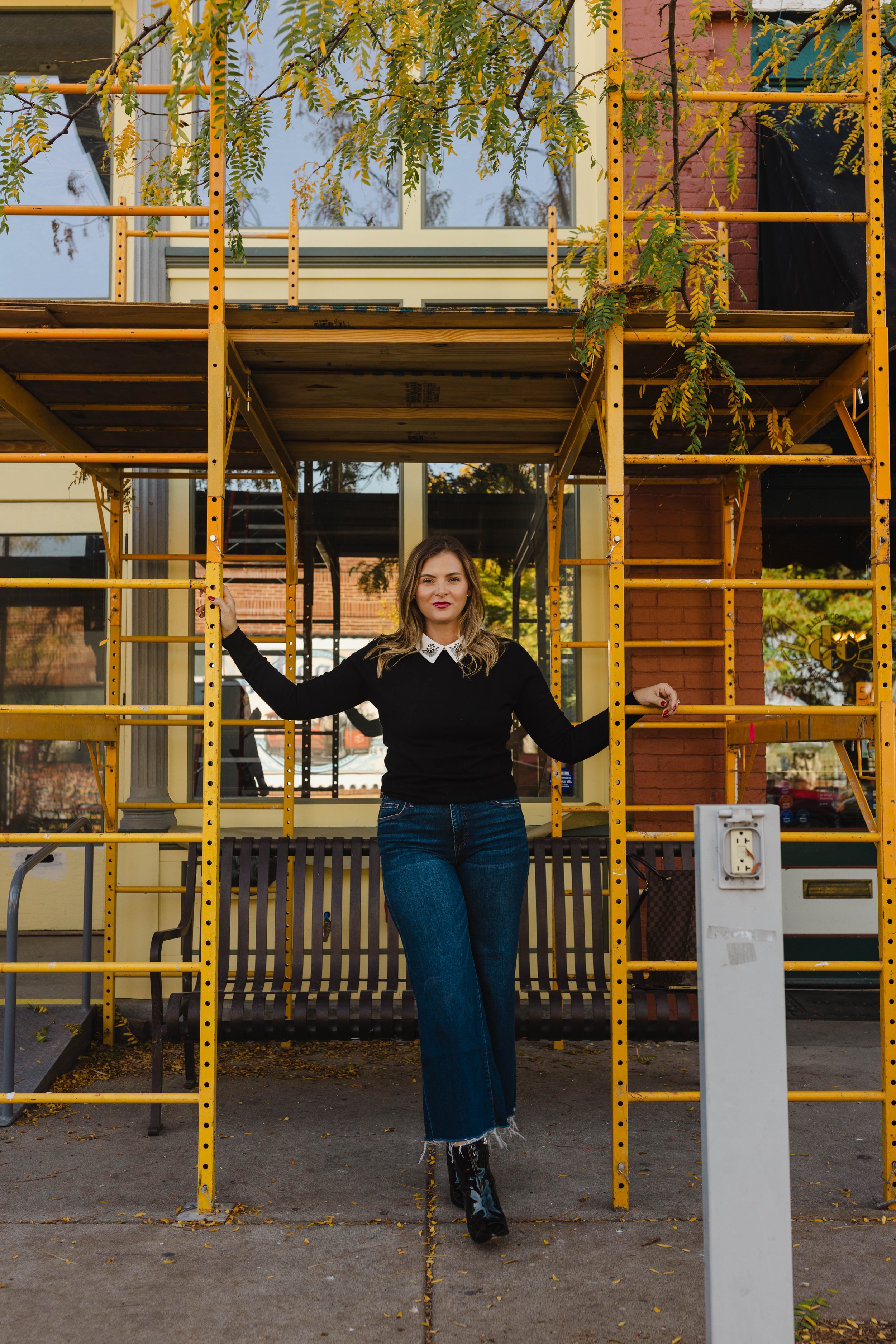 A woman with light brown hair and red lipstick standing in front of a yellow construction scaffold with green leafy branches and a building with large windows behind her.