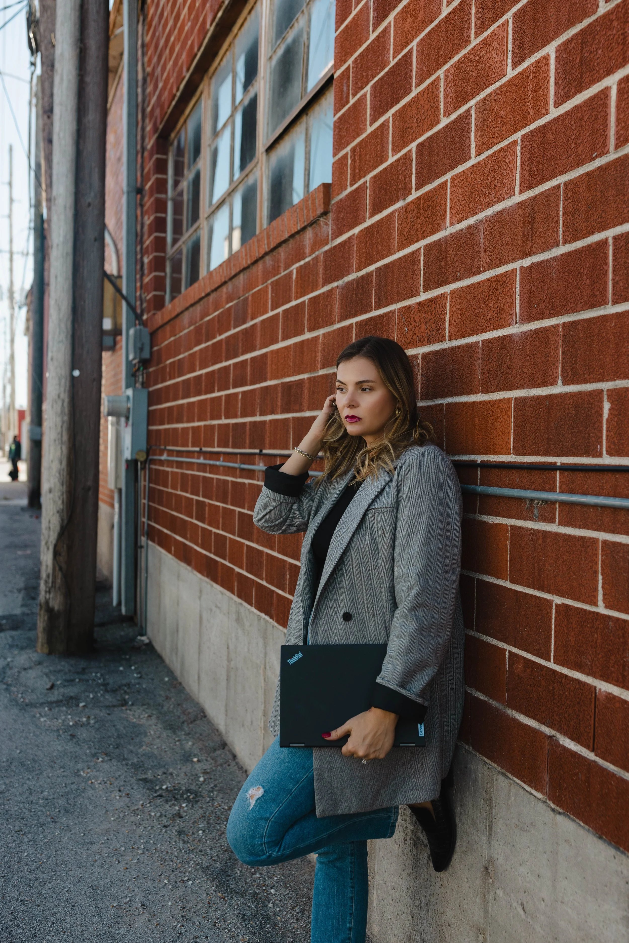 A woman wearing a gray blazer, black top, and ripped blue jeans leaning against a brick wall while holding a closed black laptop, with her right hand touching her ear, on a city sidewalk.