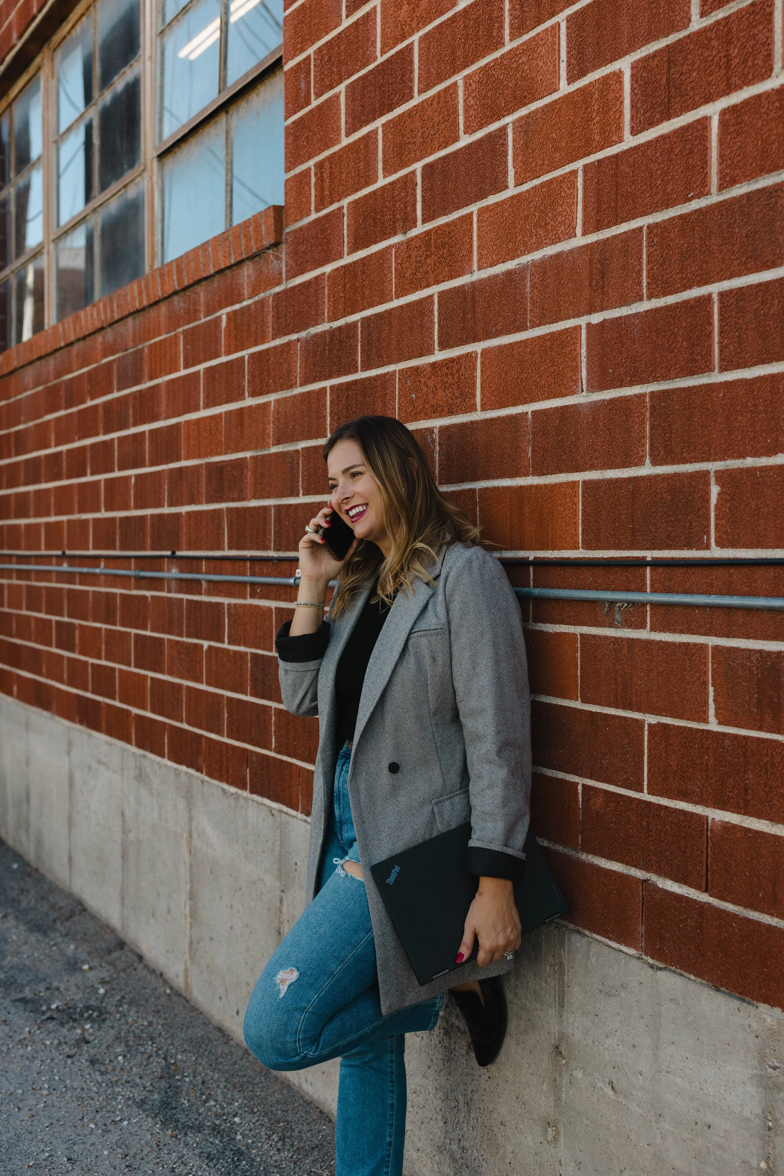 A woman standing against a red brick wall, talking on her cellphone and holding a closed laptop, smiling and dressed in a gray coat and ripped jeans.